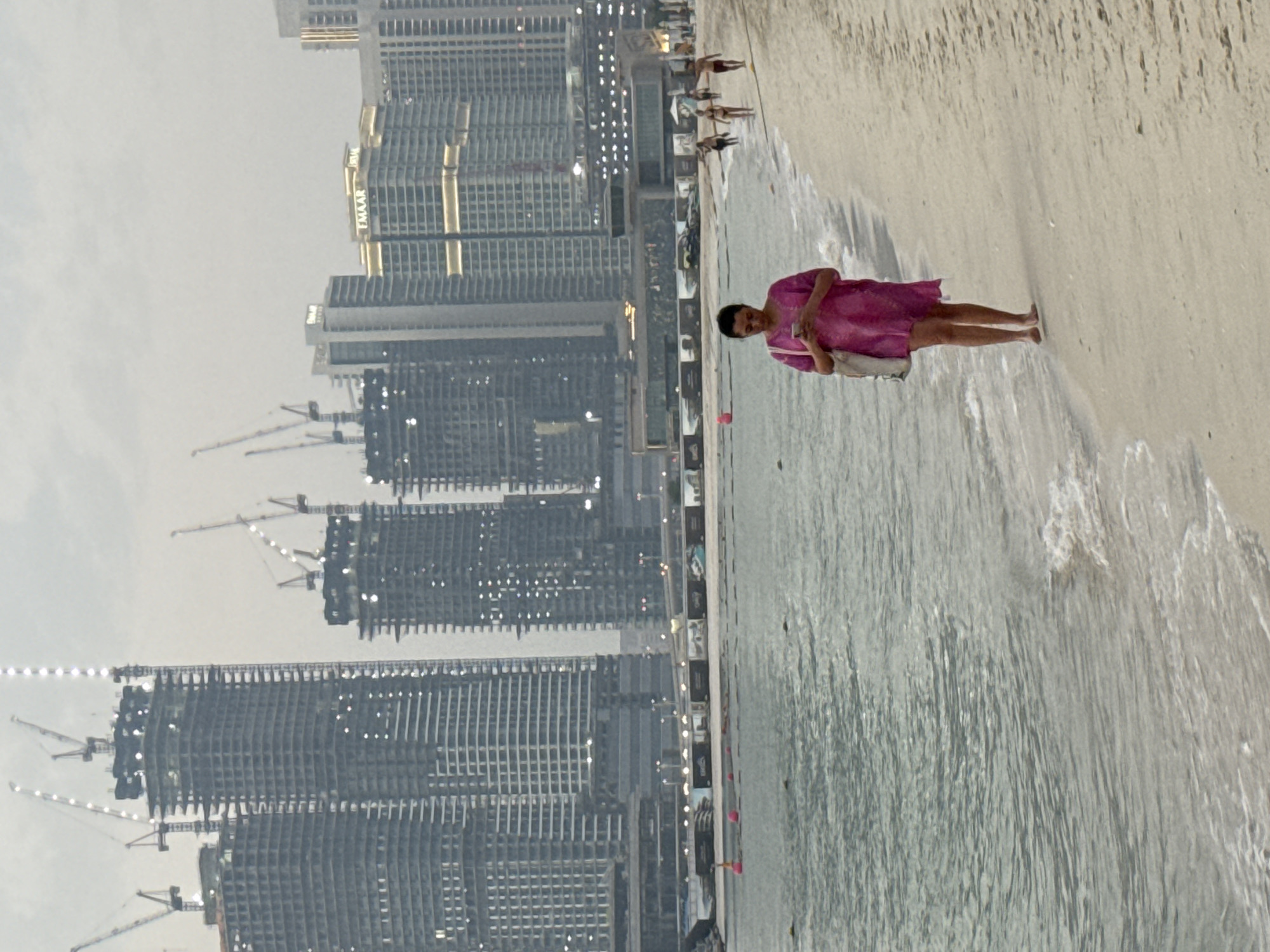 A person wearing a pink dress stands on a sandy beach with gentle waves lapping at their feet. The person is holding a light-colored bag over their shoulder. In the background, a line of modern skyscrapers rises against a hazy sky, some appearing to be under construction with cranes visible. The scene suggests an urban coastal environment. The overall lighting indicates it could be daytime, possibly overcast or early morning/late afternoon due to the softness of the light. The presence of the beach and the water contrasts with the dense urban development behind it. There is no visible text in the image.