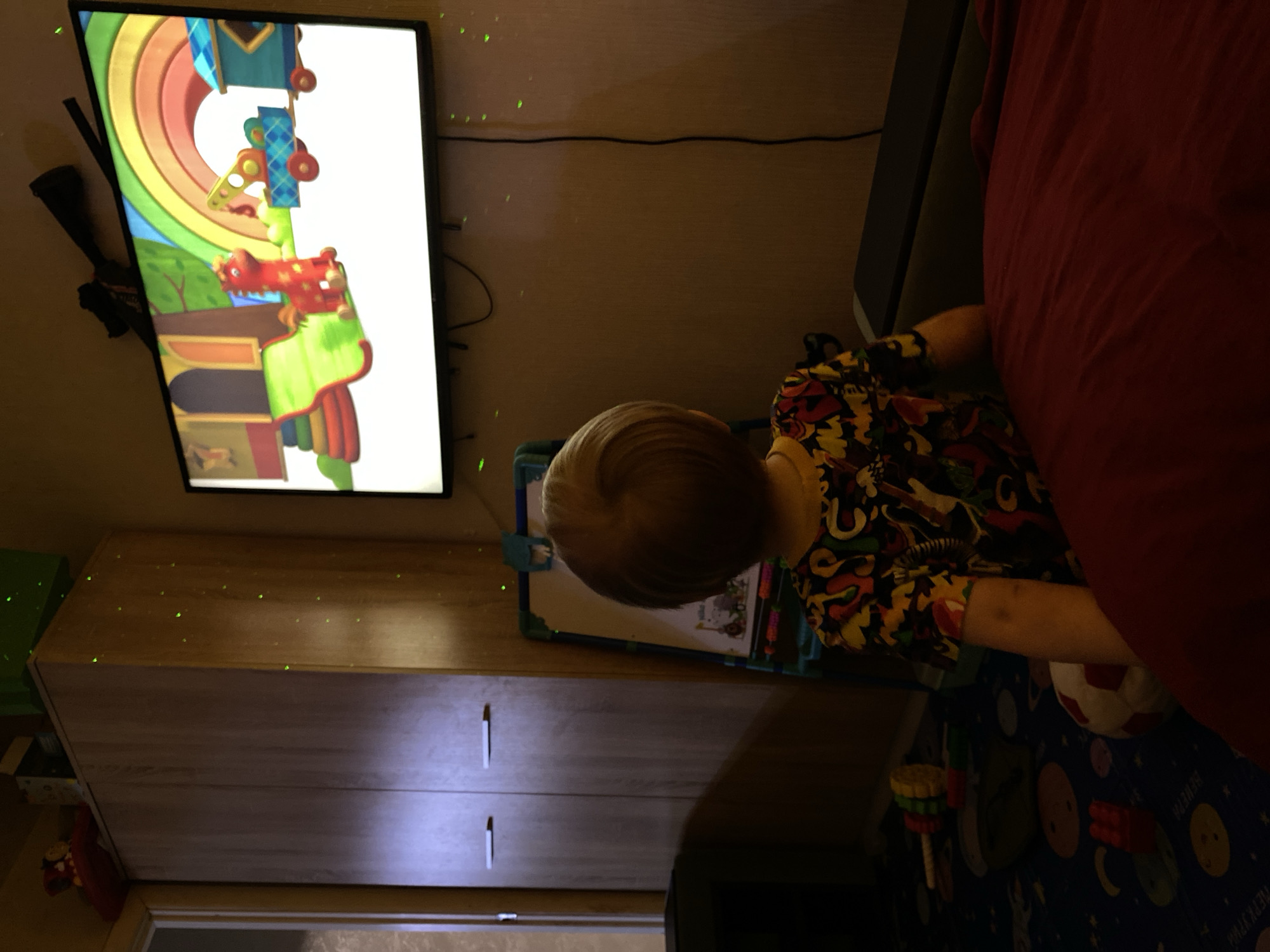 A young child is seated, facing a television screen displaying animated content. The child is wearing a multicolored shirt. The television is mounted on a wall above a wooden cabinet. The surrounding environment includes a red duvet or blanket, a child's drawing board, and a bed. The scene appears to be indoors, possibly a child's bedroom. There is no information in the image to suggest a specific location in Votkinsk, Russia.