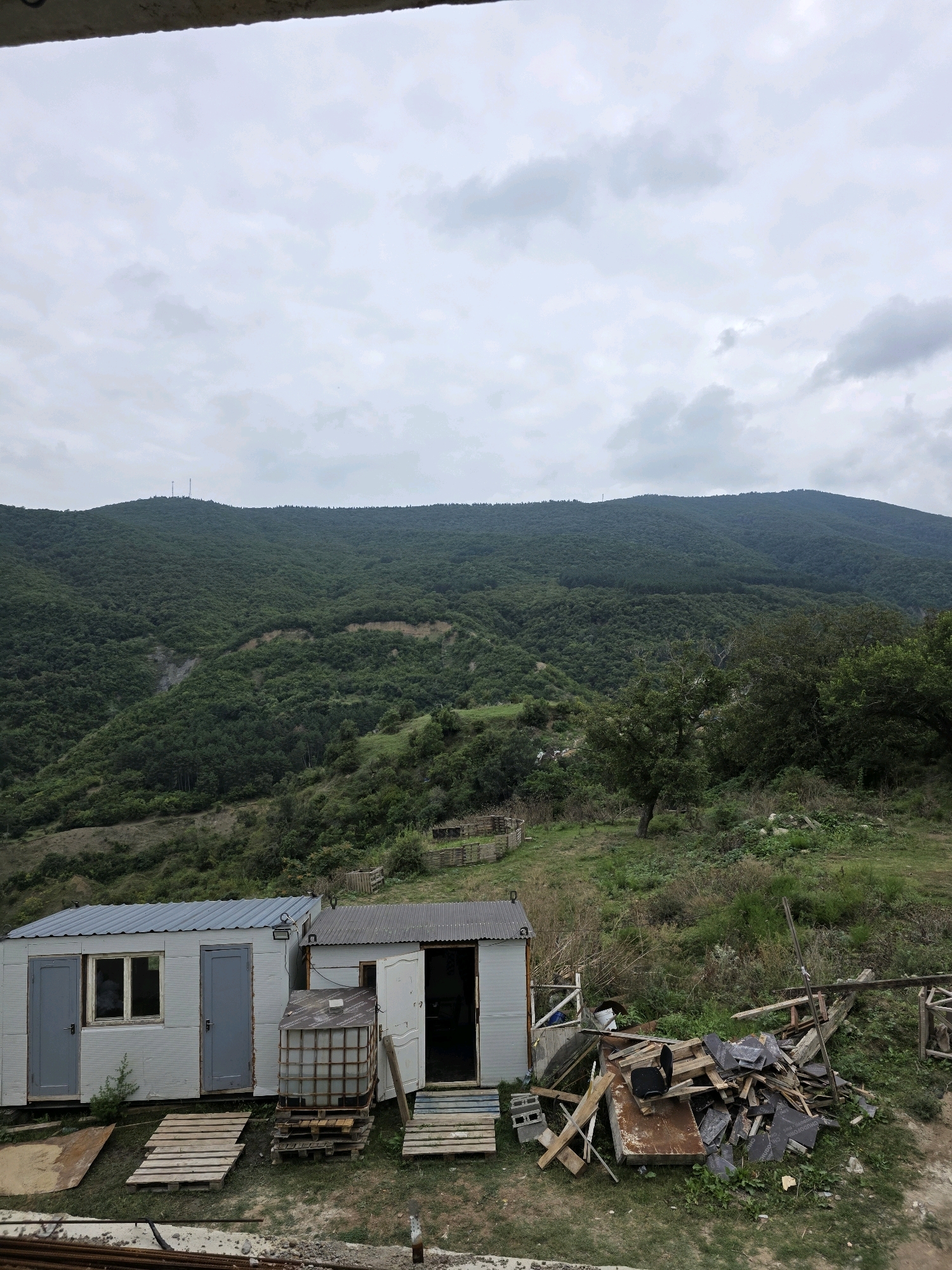 The image depicts a view from a high vantage point, looking out over a hilly, forested landscape, likely on the outskirts of Tbilisi, Georgia. In the foreground, two prefabricated structures, possibly temporary housing or storage units, are visible. The structure on the left has two gray doors and one window, while the structure on the right has an open doorway revealing the interior. In front of these structures, there is a pile of lumber and debris, as well as several wooden pallets. A large, transparent water tank on a pallet sits between the two structures. The background is dominated by a large, densely forested mountain range under a cloudy sky. Two communication towers are visible on the crest of the mountain. The overall scene suggests a somewhat undeveloped or rural area, possibly a construction site or a remote dwelling. There are no people visible in the image.