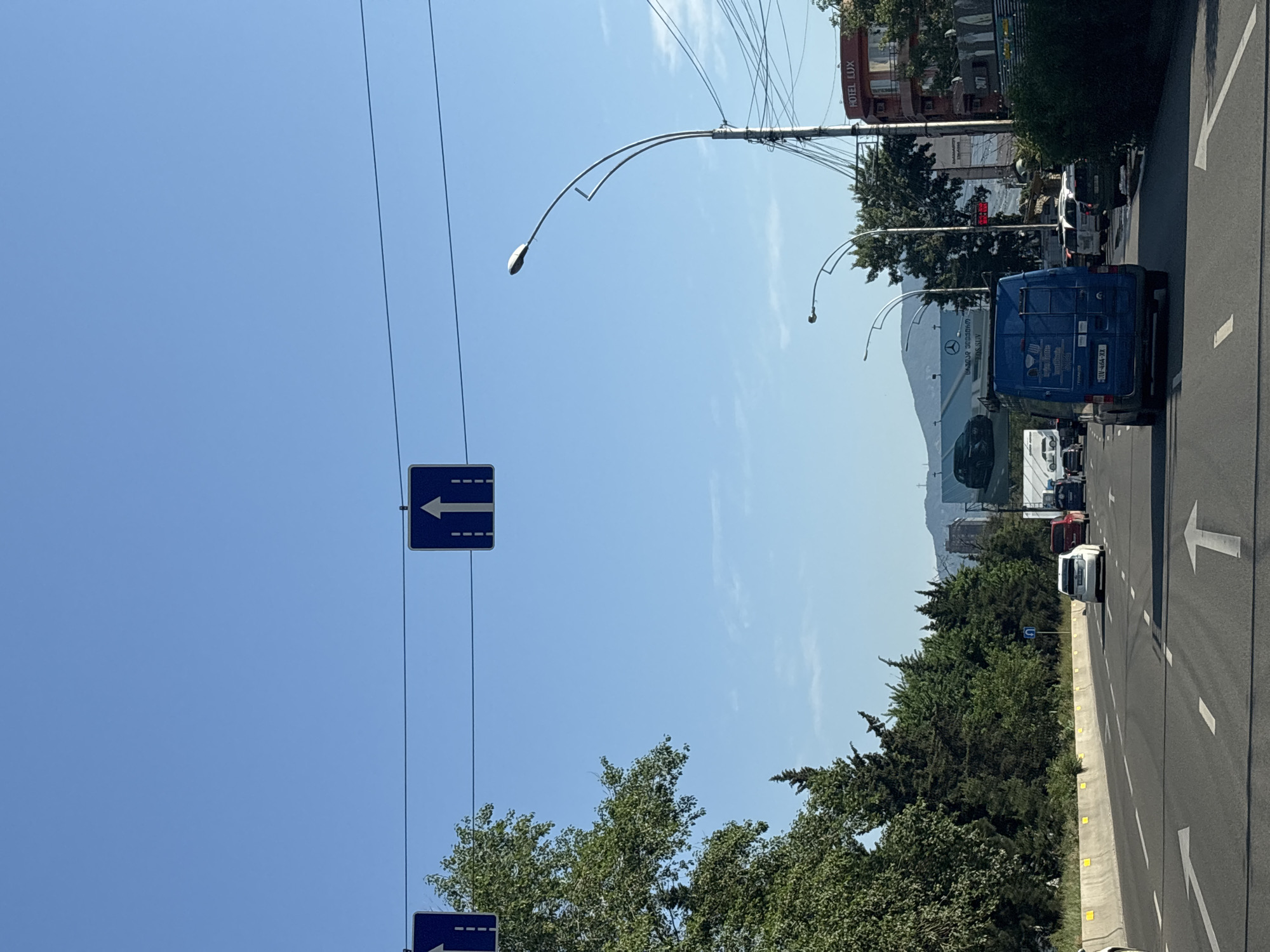 The image captures a street scene in Tbilisi, Georgia, on a clear, sunny day. The perspective is from a vehicle driving on a multi-lane road. Overhead traffic signs with white arrows pointing straight ahead are visible, indicating traffic flow. To the left, lush green trees line the roadside.
Several vehicles are on the road, including a blue van with license plate "JN-464-WX", a white car, and other cars in the distance. A concrete barrier separates the lanes of traffic from the area with trees. In the background, buildings, including a red-tiled building with a "HOTEL LUX" sign, are visible. Hills or mountains form the distant backdrop under a clear blue sky. Power lines and a street lamp are also present in the scene. The overall atmosphere is that of a typical urban environment during daylight hours.