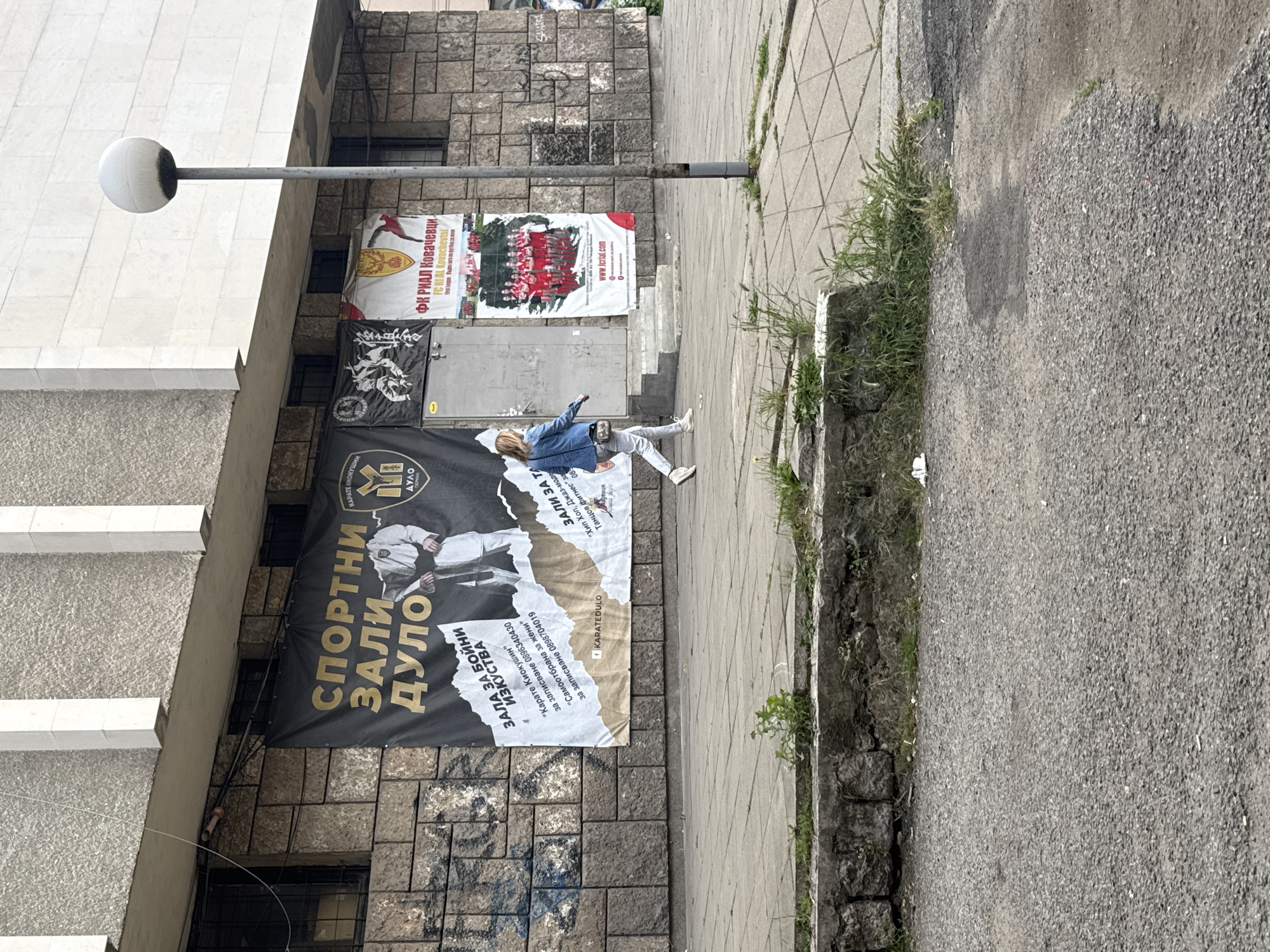 A street scene in Pernik, Bulgaria, features a sidewalk with a banner advertising a martial arts club and a poster for a football team. In the foreground, the pavement is cracked and overgrown with grass. A woman in a denim jacket and light-colored pants walks across the sidewalk. Behind her, a large black banner displays the text "СПОРТНИ ЗАЛИ ДУЛО" (Sport Halls Dulo) and images of karate practitioners. To the right, a smaller poster shows a group of young women in red uniforms, identified as "ФК РИЛ КОВАЧЕВИЦИ" (FC RIL KOVACHEVICSI), with the text "First League". A lamppost stands on the right side of the image. The building in the background has a stone facade.