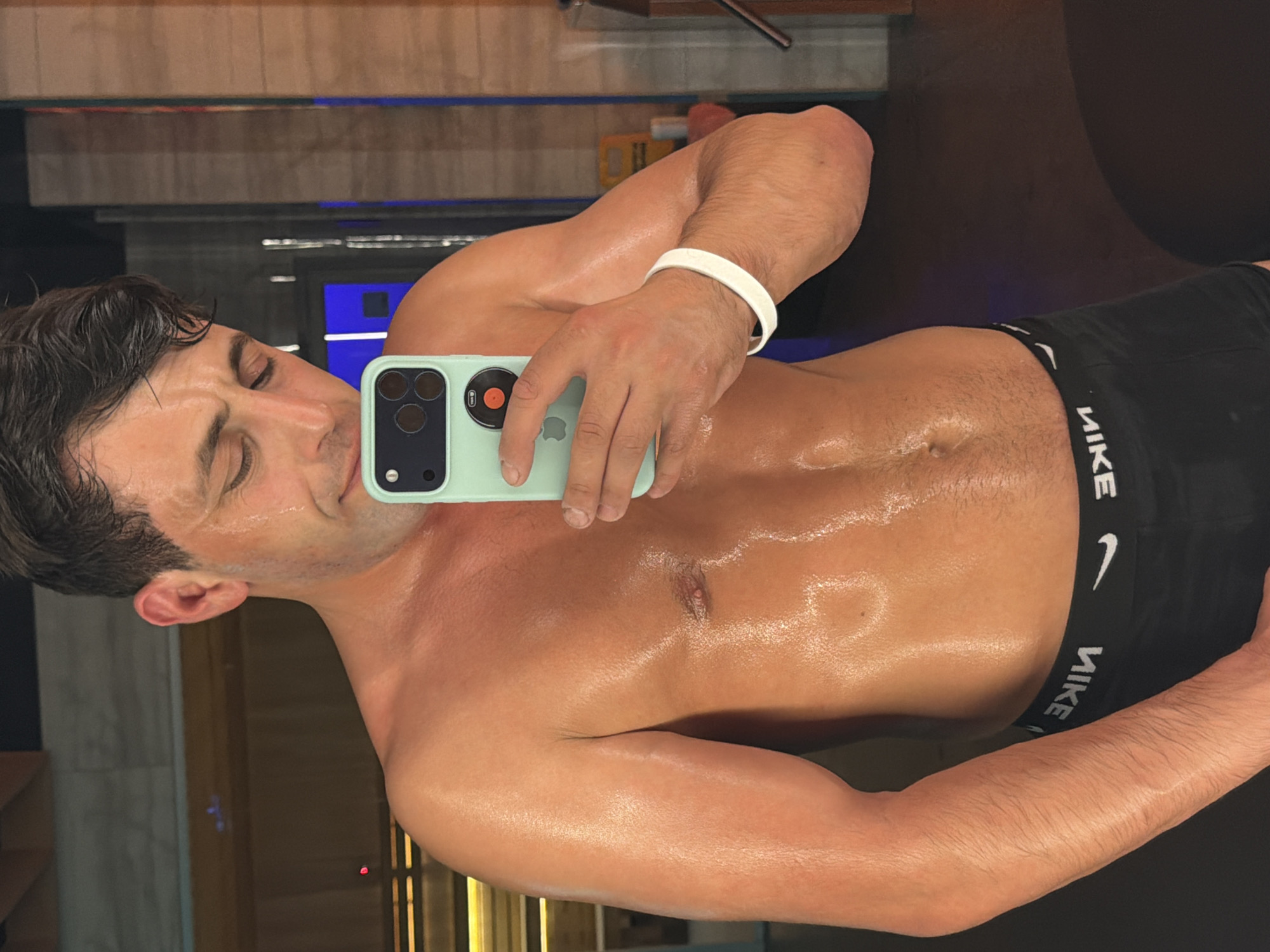 A man, appearing to be in his 20s or 30s, is captured in a mirror selfie in an indoor setting in Krasnoyarsk, Russia. His bare torso is visibly glistening with sweat or oil, highlighting his physique. He has dark, wavy hair and a light to medium complexion. His eyes are closed or cast downwards, and a relaxed, possibly self-assured expression plays on his face.
He is holding a light mint green or teal-colored smartphone, which appears to be an iPhone (the Apple logo is partially visible upside down on the back), with a distinct camera module featuring multiple lenses. A white wristband is worn on his right wrist. The man is dressed in black boxer briefs, the waistband of which prominently displays the white text "NIKE" along with the recognizable Nike swoosh logo. The background is dark and indistinct, showing some blurry reflections and hints of light sources, suggesting a private or semi-private indoor environment like a gym changing room, sauna, or bathroom.
