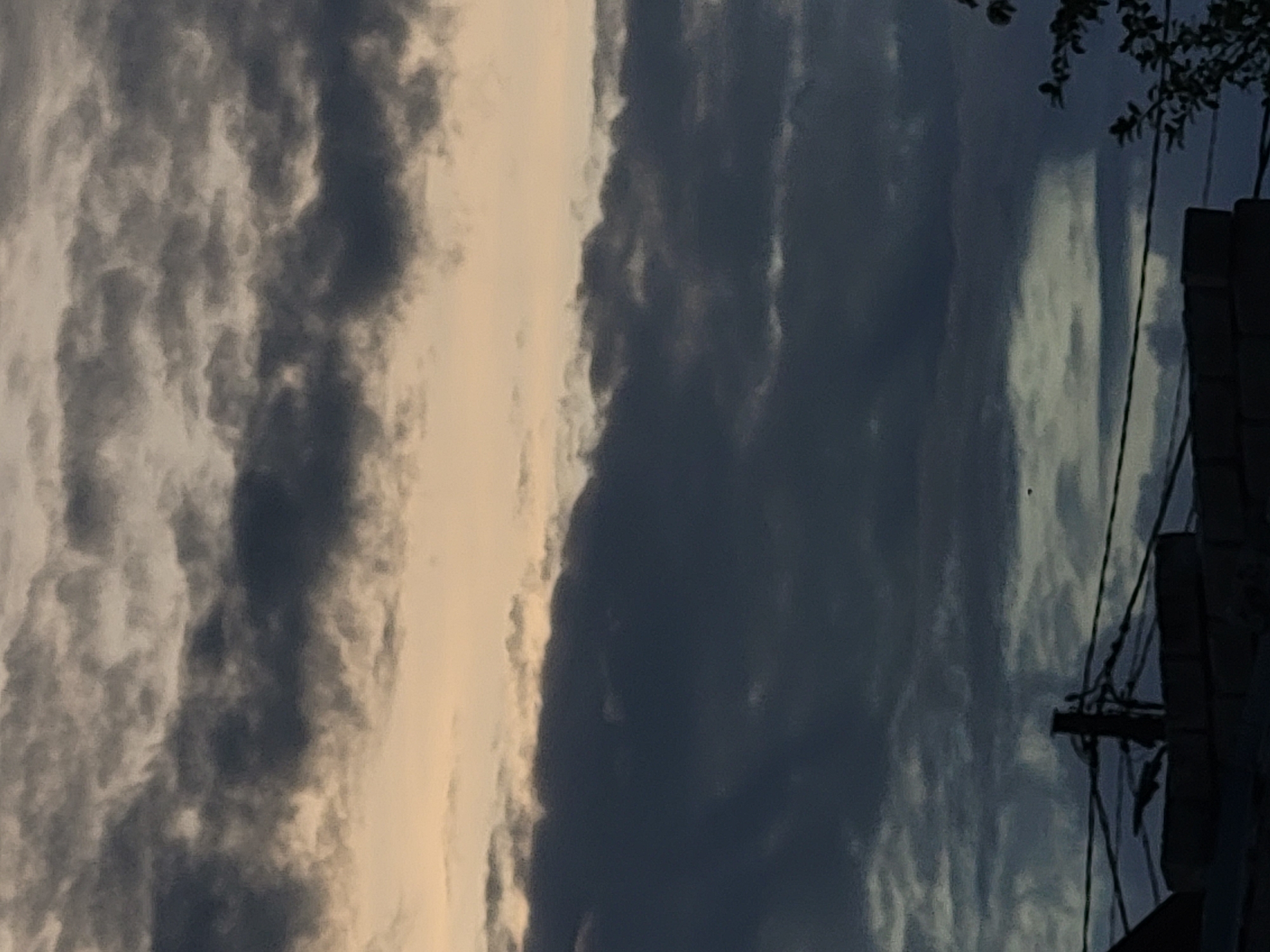 A wide shot captures a dramatic sky filled with dark, heavy clouds. The clouds are layered, with darker, more dense formations in the foreground and lighter, wispy clouds in the background. The colors range from deep grays and blues to lighter shades of beige and pale yellow, suggesting either an impending storm or the aftermath of one. In the lower portion of the frame, silhouetted against the sky, are the tops of buildings and a complex network of wires and a wooden post, indicative of an urban environment. A few sparse tree branches are visible on the far right, also silhouetted. The overall impression is one of overcast, possibly moody weather.