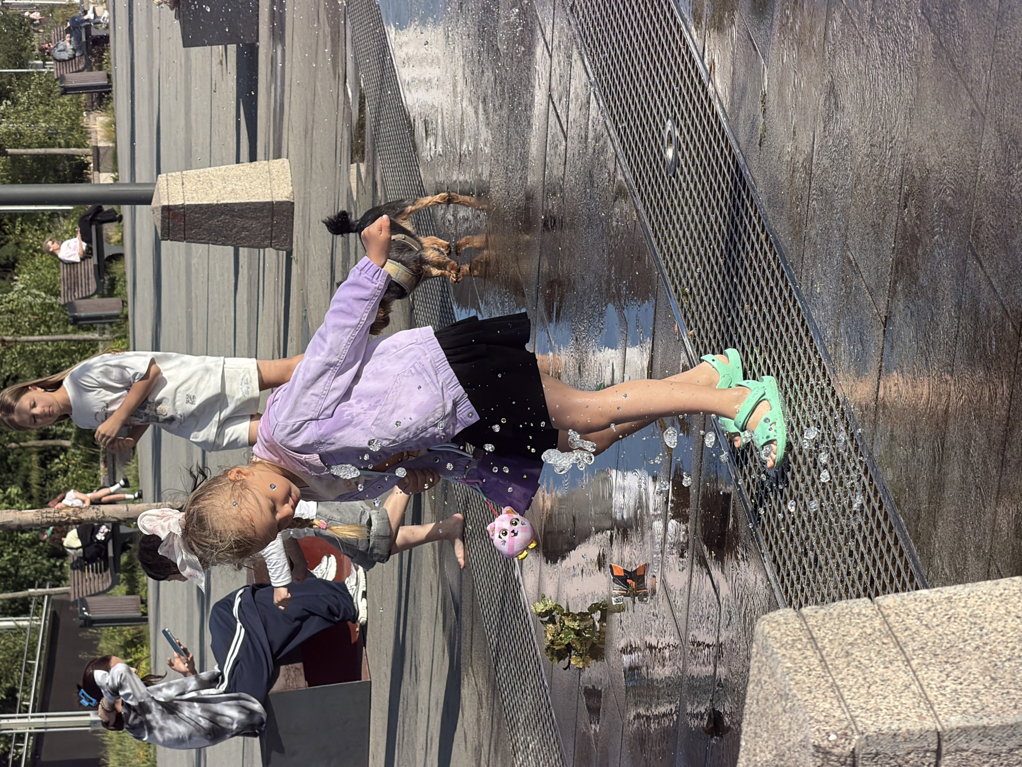 This media file shows a group of people, including children and at least one dog, interacting with a water feature. The scene appears to be an outdoor public space, possibly a park or plaza, with paved ground, some benches, and trees visible in the background. The time of day seems to be daytime, with bright sunlight casting shadows and creating highlights on the wet surfaces. The weather appears to be warm and sunny, as people are dressed in light clothing and engaging with water.
The main focus is on a young girl in a purple jacket and black skirt, playing in the water. She is wearing light green sandals. Another child, a girl with blonde hair and a white t-shirt, is standing nearby. A dog, which appears to be a terrier mix, is also in the water, being interacted with by one of the children. Other people are visible in the background, some sitting on benches and others standing.
Notable details include the way the water is spraying and reflecting the sunlight, creating a dynamic and playful atmosphere. There are also some decorative elements visible, such as a small plush toy that looks like a pink cat, and a decorative butterfly. The ground is wet and appears to be made of dark paving stones or tiles, with a metal grate running along one side of the water feature.
The overall mood seems to be joyful and relaxed, with the children clearly enjoying their time playing in the water. There is no visible text in the image.
