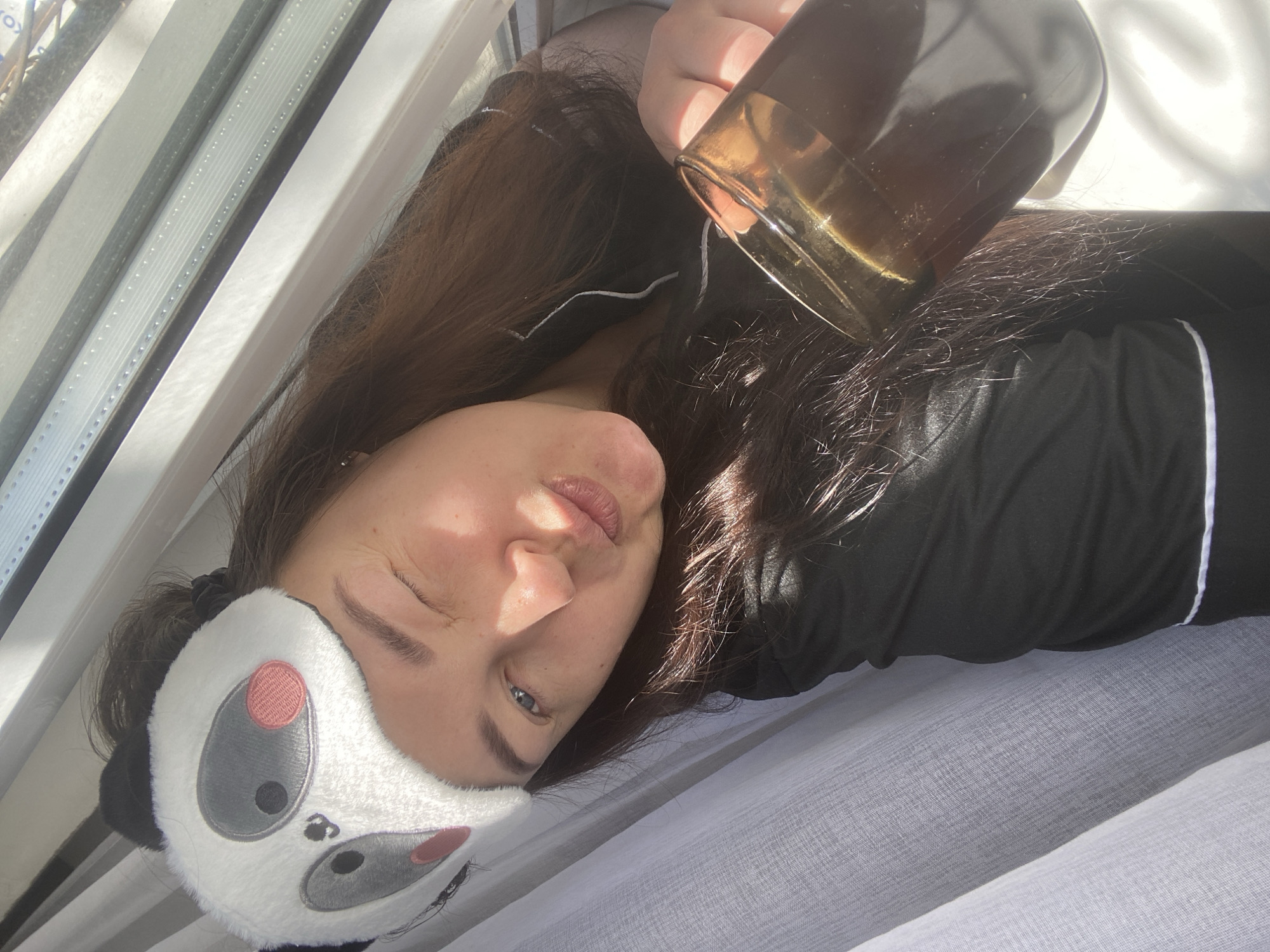 The image, set in Sochi, Russia, captures a close-up of a woman lying down indoors, bathed in bright natural sunlight. She is positioned diagonally in the frame, wearing a black long-sleeved top with white piping, suggestive of pajamas. Her dark brown hair is spread around her head and shoulders.
A distinctive panda-themed sleep mask, predominantly white with black and pink details, is pushed up onto her forehead and hair, revealing her face. One of her eyes is partially open and squinting, while the other appears closed or heavily squinting, possibly due to the direct sunlight. Her expression conveys a sense of relaxation, perhaps a gentle awakening, or being slightly bothered by the intense light.
She holds a clear glass with a dark liquid, likely coffee or tea, in her right hand, visible towards the top right of the frame. The glass has a reflective, possibly gold-toned, rim.
The background features a window with horizontal blinds or louvers on the left, allowing strong sunlight to stream into the room. Sheer white curtains are visible along the bottom and right edges of the frame, contributing to the bright, sun-drenched atmosphere. The interplay of light and shadow is prominent on her face and the surrounding elements, indicating a sunny day, likely morning or early afternoon. There is no visible text in the image.