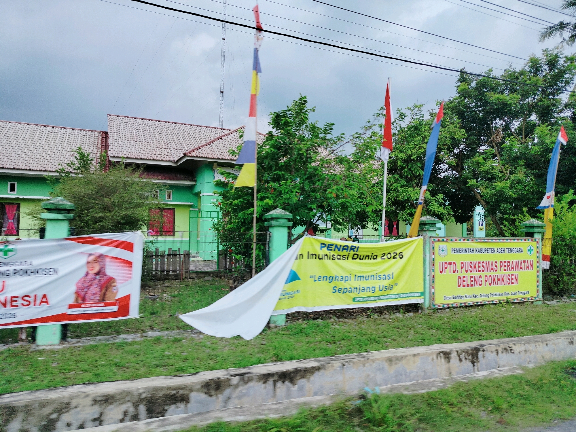 The image shows a street view in Beriringnaru, Indonesia, with a green building and several banners and flags. The building appears to be a health facility, as indicated by the banners. One banner on the left is white with red trim, featuring a picture of a woman in a headscarf. Another banner, yellow, displays text in Indonesian related to immunization and a date, "2026". A third banner on the right has a green and red border and lists the name of a government health department, "UPTD. PUSKESMAS PERAWATAN DELENG POKHKISEN". Several colorful flags, including red and white Indonesian flags, are also visible. The scene is set against a backdrop of trees and an overcast sky, with power lines overhead. The street in front of the building has a concrete curb and grassy areas.