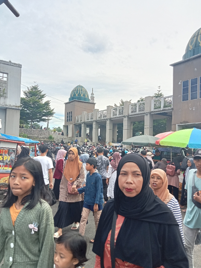 This daytime image captures a crowded street scene in Singaparna, Indonesia, beneath an overcast sky. A large number of people, including adults and children, fill the street, indicating a busy gathering, possibly a market day or community event.
In the foreground, a woman wearing a black hijab and a black garment with red accents looks directly at the camera, her lips a notable red. To her left, a young girl with dark hair and a green cardigan also faces the camera. A smaller child's head is visible at the bottom center of the frame. Behind these individuals, the street is densely populated with more people walking and interacting. Many women in the crowd are wearing hijabs, and a woman in a brown hijab and striped top holds a boy's hand.
The backdrop features a prominent mosque with a distinctive green dome and a slender minaret, characteristic of Southeast Asian Islamic architecture. The mosque structure includes an elevated walkway or bridge. To its right, an adjacent building has blue-tinted windows, while to its left, another building appears to be in an unfinished state with exposed concrete.
Evidence of commercial activity is present: a red and white vendor cart is partially visible on the left, displaying fragmented text "HU SP." On the right side of the street, colorful market umbrellas suggest additional stalls or vendors. Further into the background, numerous motorbikes are seen amidst the crowd, indicating a bustling urban or town environment. The overall impression is one of lively community activity.