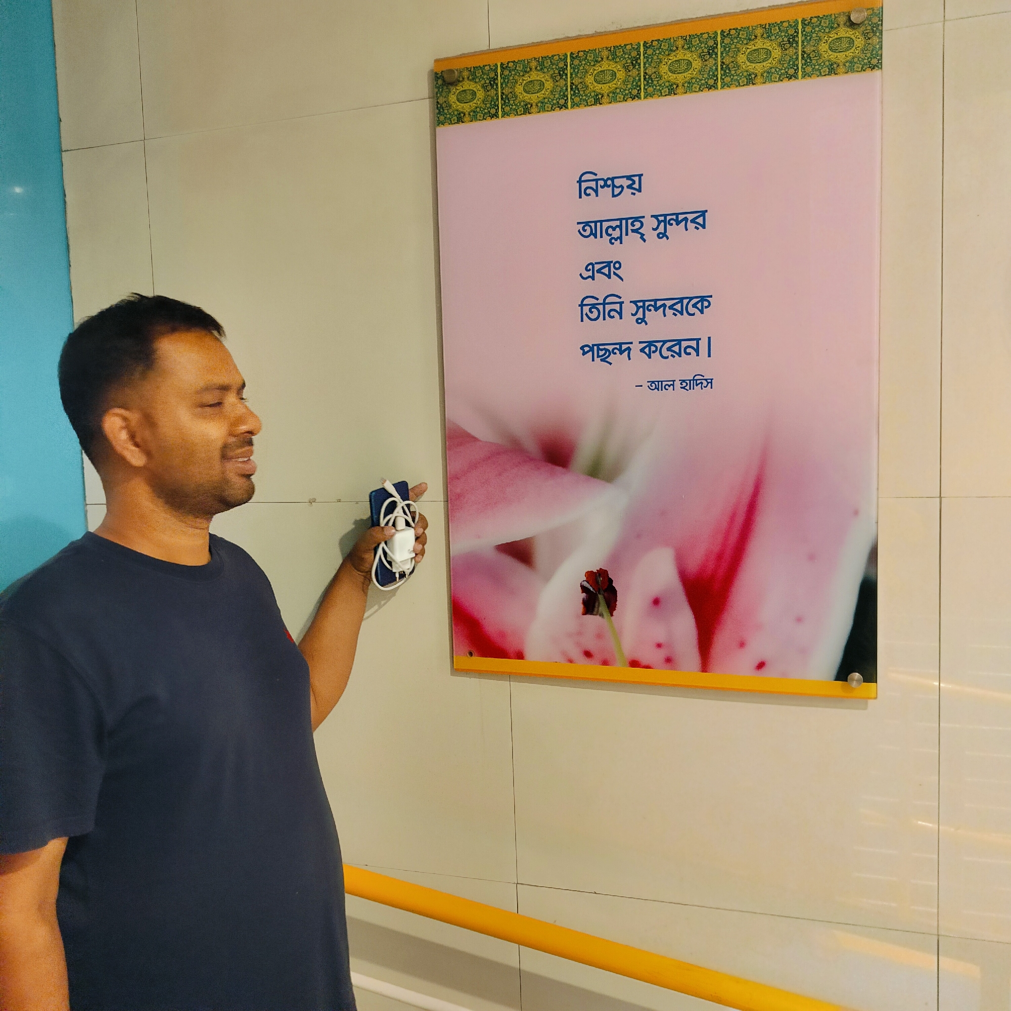 The image captures a man in Dhaka, Bangladesh, standing next to a sign written in Bengali. He is middle-aged, with dark hair and skin, and is wearing a navy blue t-shirt. In his right hand, he holds a smartphone connected to a charger. He appears to be looking at the sign.
The sign is framed in yellow and displays Bengali script in blue font on a light pink background. An image of a flower is also visible on the sign. The top border of the sign features a decorative pattern in green and gold. The sign is mounted on a white tiled wall. A yellow handrail is visible along the wall. The overall lighting of the scene appears bright.