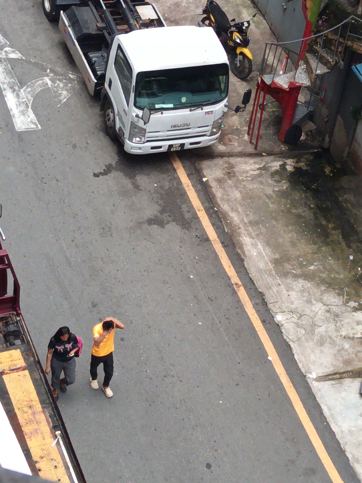 The image captures an aerial view of a street scene in Kuala Lumpur, Malaysia. In the center, a white Isuzu truck is parked. To the right of the truck, a yellow and black motorcycle is parked. In the lower portion of the image, two individuals are walking on the street. One is a woman wearing a dark shirt with a bow pattern and gray pants, carrying a pink backpack. The other is a man in an orange t-shirt and black pants. The street is paved with asphalt and marked with a yellow line. There are also some concrete areas and debris visible on the sides of the street.