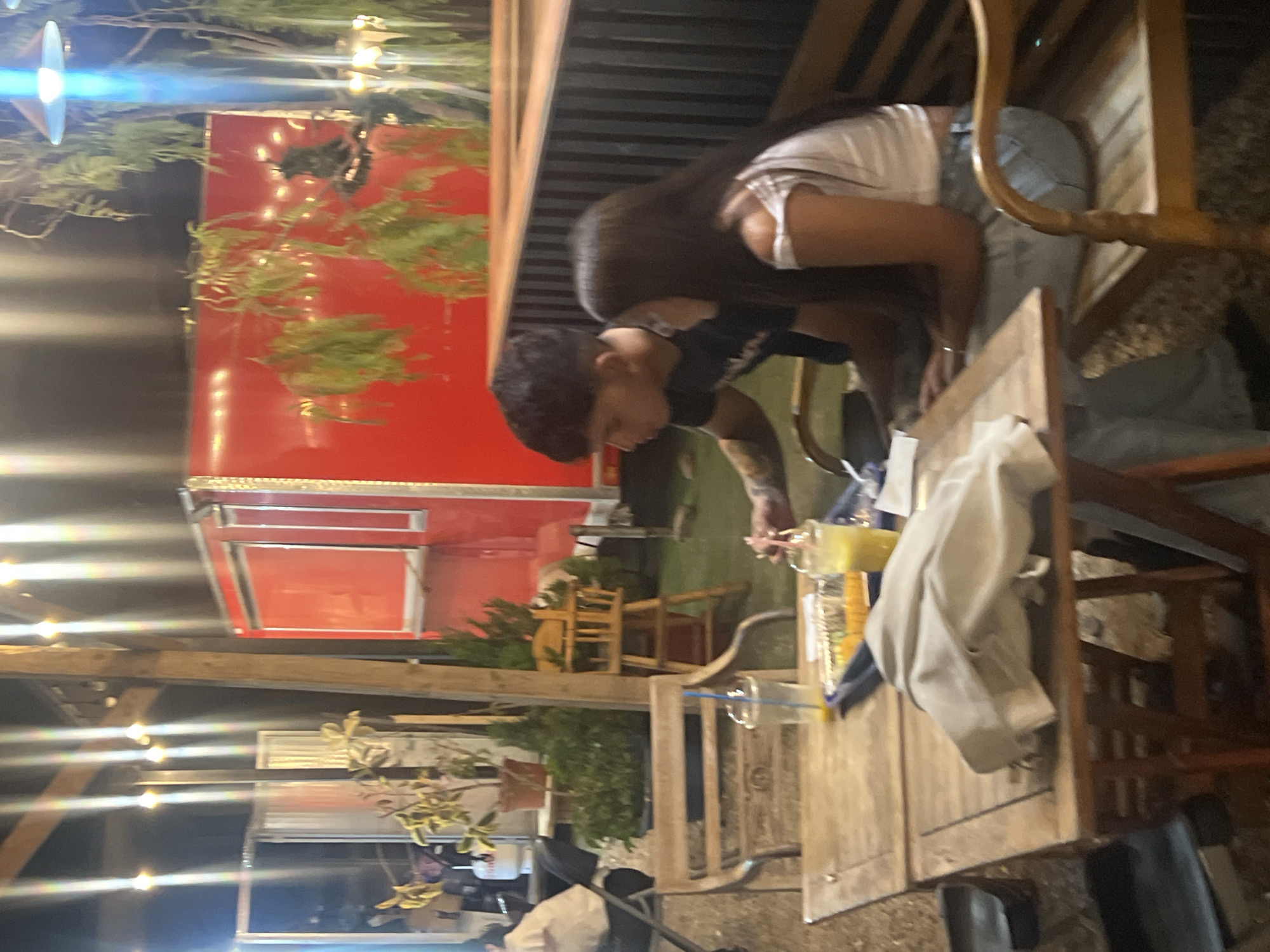 The image depicts a nighttime scene in what appears to be an outdoor dining area, possibly in Arica, Chile, although explicit city evidence is not present. The main subjects are a couple seated at a wooden table. The male figure has dark hair and is wearing a dark shirt and a face mask partially pulled down. The female figure has long dark hair and wears a white shirt. They seem to be leaning towards each other.
On the table, there's a glass of a yellowish beverage, possibly juice or a cocktail. A tan jacket or coat is draped over the back of a chair. The table itself is made of light-colored wood.
In the background, there's a bright red food truck or container adorned with green foliage. String lights are visible above, suggesting a casual, outdoor setting. A scattering of other chairs and tables implies a larger dining area. The general environment appears to be a restaurant or food court setting.