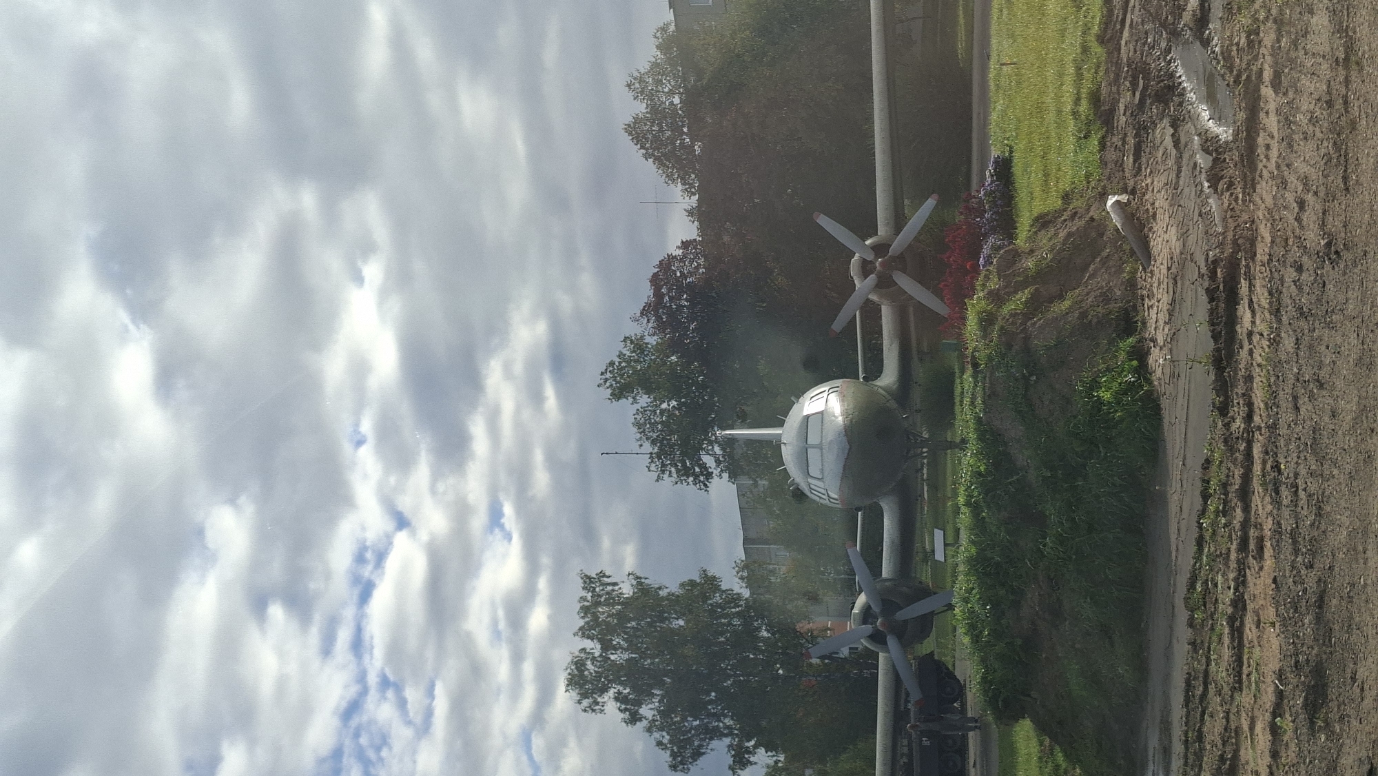 An abandoned airplane with two propellers sits in a grassy area. The airplane is painted a dull green and has a silver nose. It is surrounded by trees and a building in the background. In the foreground, there is a dirt path with some puddles. The sky is cloudy and overcast. There is no indication of the location being in the city of Бягомльскі сельскі Савет, Belarus.