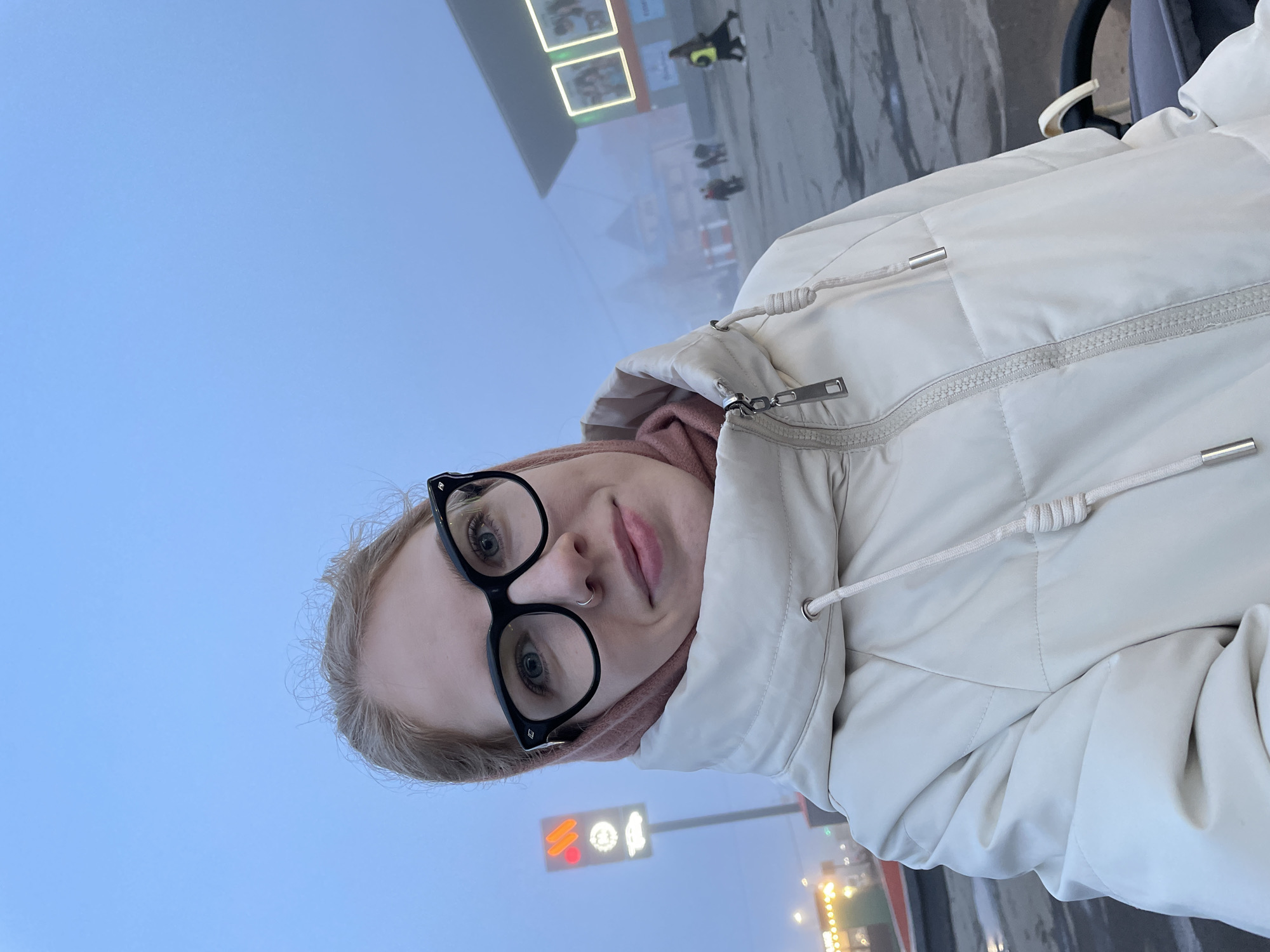 A woman with light hair and large black glasses is taking a selfie. She is wearing a white puffer jacket. A traffic light with a red signal is visible behind her. In the background are buildings and people, partially obscured by fog. The location is Izhevsk, Russia.