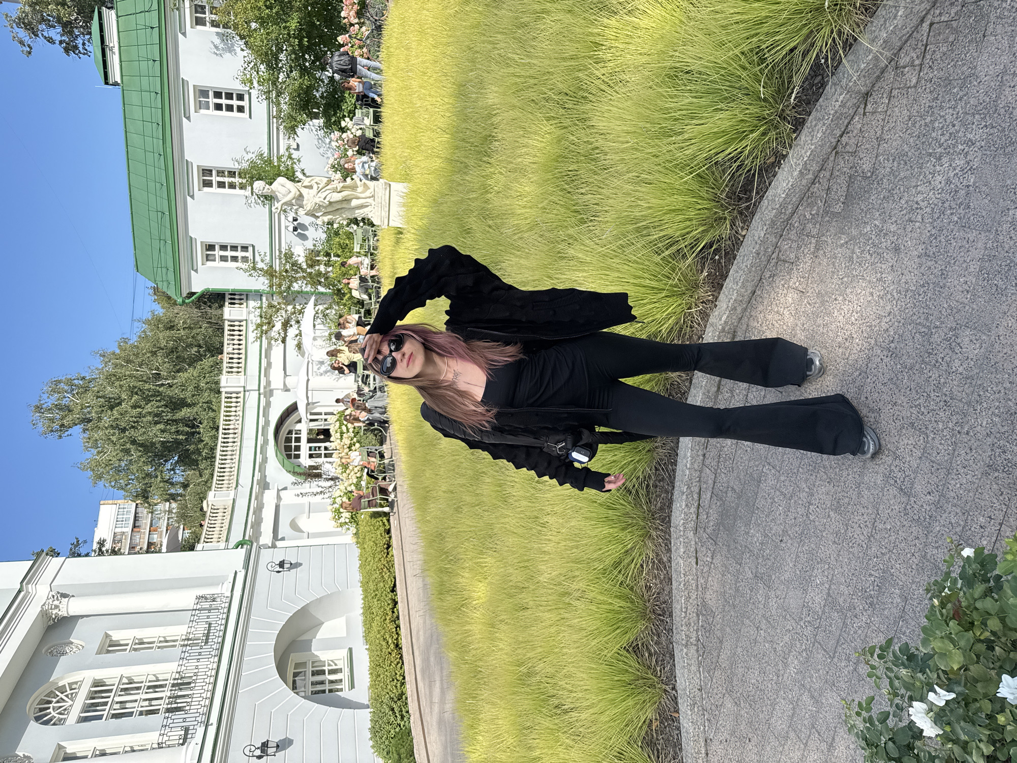 A woman stands in a courtyard with a paved walkway and manicured grass in front of a light blue and green building. The woman is wearing black flared pants, a black tank top, and a black zip-up jacket with a textured pattern. She has dark sunglasses on and her left hand is on her forehead. She is looking towards the camera. In the background, people are seated at tables in front of the building, and a white statue stands in the grass. The building appears to be a historical structure with classical architectural elements. The setting is likely a public garden or park. The location is the Museum-Estate of Ostankino in Moscow, Russia.