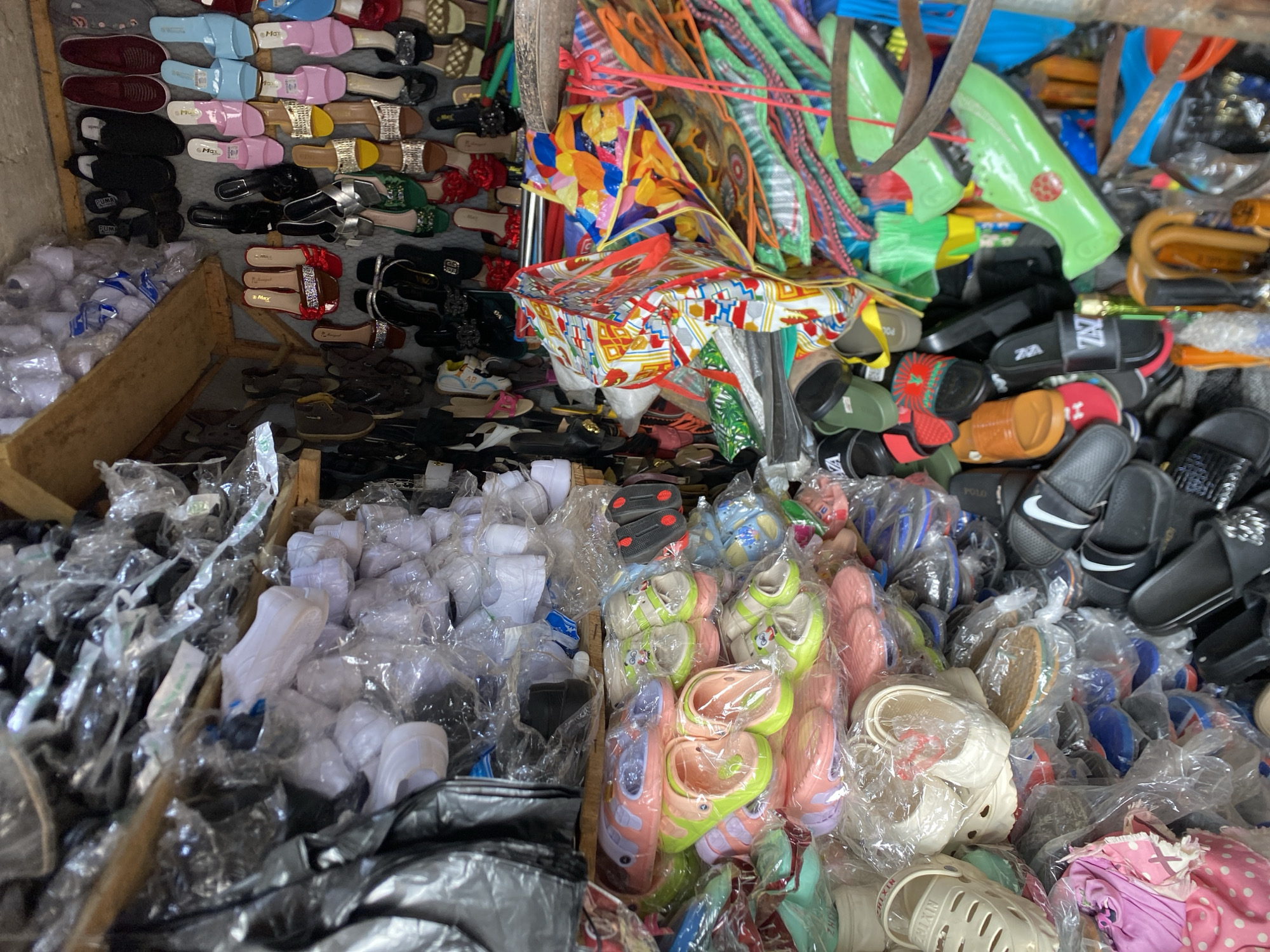 The image depicts a crowded market stall in Bukuru, Nigeria. The stall is filled with a variety of footwear, including sandals, slippers, and shoes, many still in plastic packaging. The footwear is arranged in different piles and displays. There are also bags, possibly containing additional merchandise, present in the scene. The stall is cluttered, with colorful fabrics, rope, and other items adding to the visual density. The lighting suggests an indoor setting, potentially within a larger market structure. There are no people visible in the immediate frame.