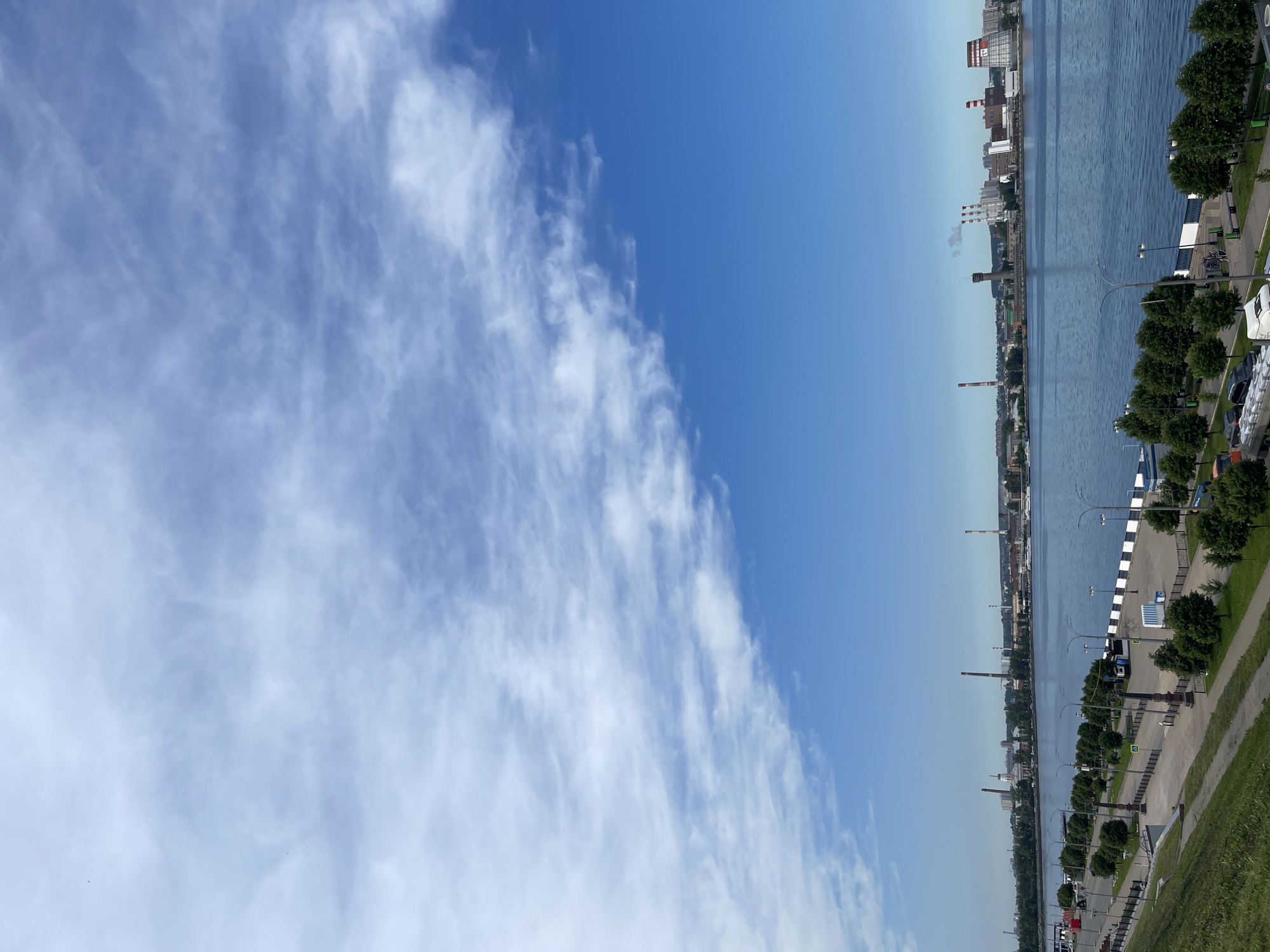 The image displays a serene urban landscape under a bright blue sky, presented as if the original vertically-oriented image has been rotated 90 degrees counter-clockwise to achieve a horizontal perspective.
The upper portion of the frame is dominated by a clear blue sky. Wispy, elongated white clouds, suggestive of cirrus formations, stretch across the left side of the sky, gradually thinning out towards the right, which remains a pristine blue.
Horizontally bisecting the mid-ground is a wide body of water, likely a river or canal, appearing calm and a deep blue under the daylight. Its surface shows subtle ripples, reflecting the sky above.
On the far bank of the water, a distant cityscape unfolds. It features a mix of buildings, including several industrial structures with tall chimneys, one of which emits a light, grayish plume of smoke. The number "12" is visible on the side of one building, and another has a distinct red and white design on its roof.
The near bank, occupying the lower third of the image, presents a paved promenade or walkway. This pathway is neatly lined with uniformly spaced green trees and streetlights. Several parked cars are visible along the promenade, which is bordered by a grassy slope rising gently towards the bottom right.
The scene captures a clear and sunny day, indicating midday or early afternoon conditions due to the strong natural light. The overall setting suggests an urban or industrial area situated alongside a major waterway. No explicit human activity or event is taking place, contributing to the tranquil atmosphere of the static landscape.