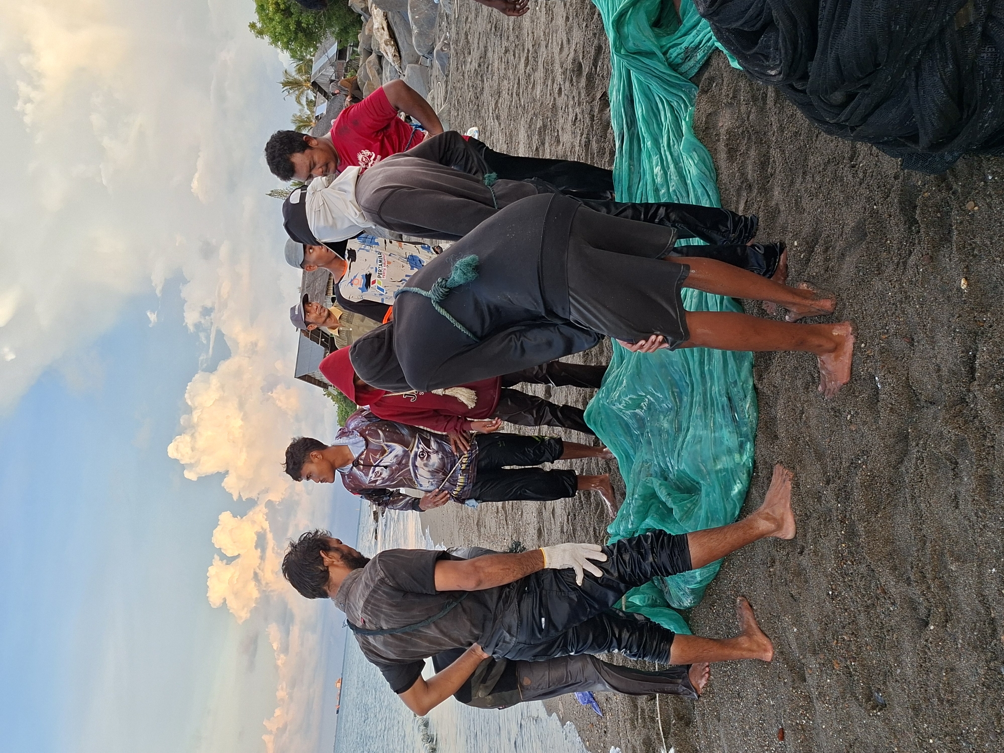 The image depicts a group of approximately ten individuals standing on a dark sandy beach. A body of water with a distant ship is visible in the background on the left. The sky is a mix of pale blue and orange-tinged clouds. The people are wearing various casual clothes, including t-shirts, hoodies, and pants. Some are barefoot, while others have their feet partially submerged in the sand. A large, green tarpaulin or net lies on the beach, with black netting visible to the right. The location is likely the beach in Lhokseumawe, Indonesia.