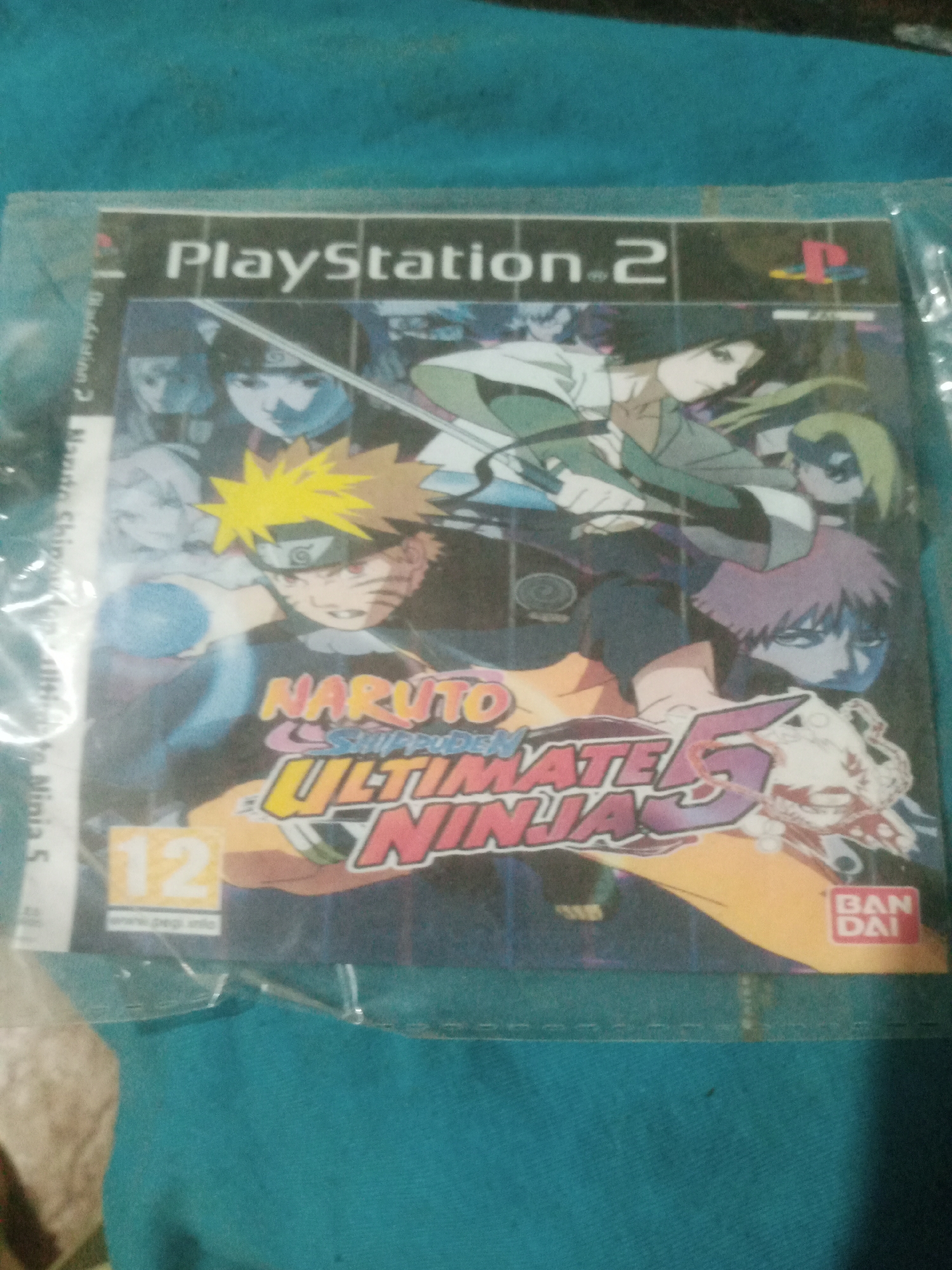 This image shows the cover of a PlayStation 2 video game titled "Naruto Shippuden: Ultimate Ninja 5". The game cover features several anime characters from the Naruto series. The cover is partially covered by clear plastic. In the top left corner, the "PlayStation 2" logo is visible. The game cover also displays a PEGI 12 rating and the Bandai logo in the bottom right corner. The image is a close-up, with the background appearing to be a turquoise fabric. The lighting suggests it was taken indoors. The location of Santa Ana, Venezuela, is not discernable from the image content itself.