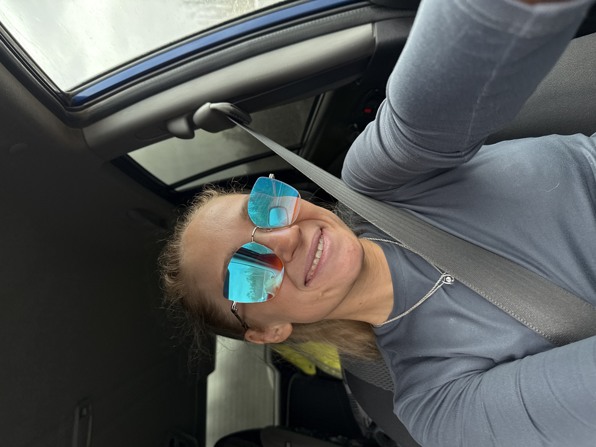 The image is a tilted, eye-level photograph taken inside a vehicle, featuring a smiling person wearing sunglasses. The main subject is an adult, appearing to be female, positioned in the foreground and wearing a long-sleeved, dark gray shirt. She has light brown/blonde hair, pulled back and visible around her ear. A silver chain necklace with a small, circular pendant is visible around her neck. Her face is turned slightly towards the camera, revealing a broad smile with visible teeth. She is wearing large, blue-tinted, mirrored sunglasses that reflect parts of the car's interior and the sky. A gray seatbelt crosses her chest, indicating she is seated in the front of the vehicle.
The setting is the interior of a modern car, specifically looking upwards towards the roof. A large sunroof is prominently featured in the upper section of the image, through which an overcast, cloudy sky is visible. The car's interior trim around the sunroof and upper windows is dark gray with some blue accents. The lighting suggests it is daytime, under cloudy weather conditions. The person's cheerful expression conveys a positive and relaxed mood. There is no visible text in the image. This scene takes place in Krasnokamsk, Russia.