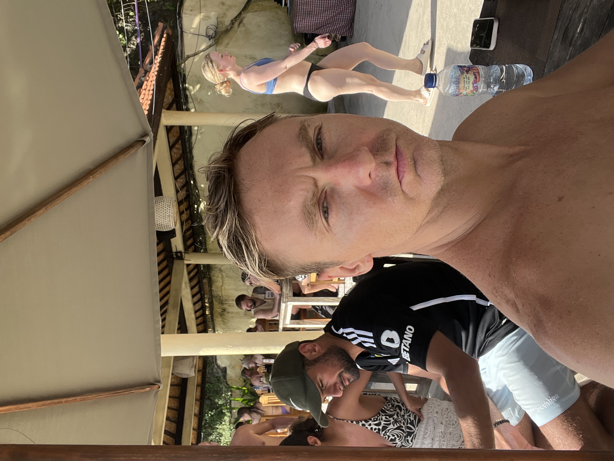 The image depicts a man in the foreground taking a selfie outdoors, likely at a social gathering or event. Behind him, there are several other people, some seated at tables under the shade of a wooden structure. A woman in a bikini is walking in the background. A bottle of water and a phone are on a table beside her. The location is estimated to be Kuta Selatan, Indonesia, given the tropical vegetation, architecture, and style of dress of the people in the image.