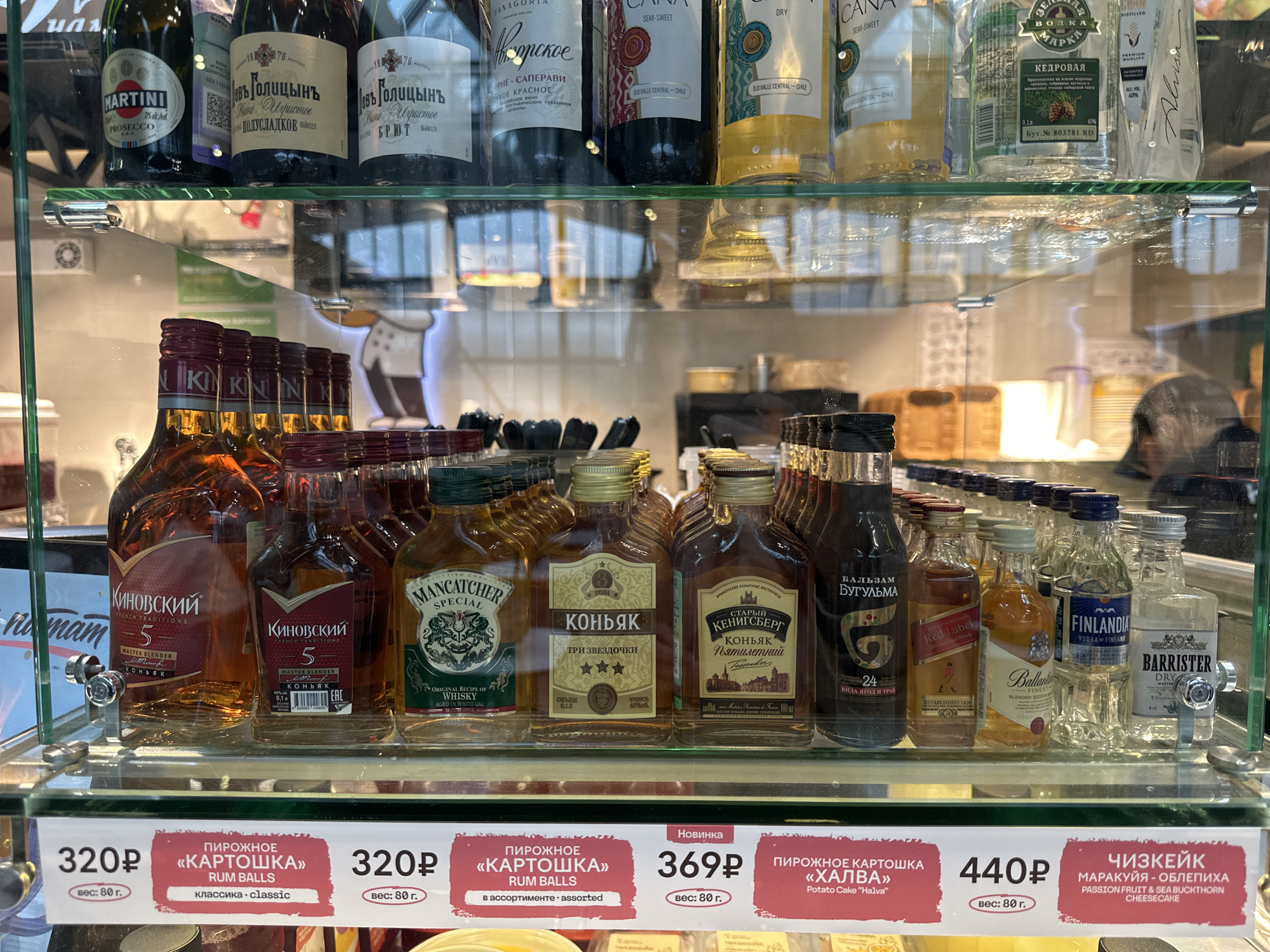 The image shows a display case containing a variety of alcoholic beverages and desserts.
On the top shelf are several bottles of wine and liquor, including Martini Prosecco, "Lev Golitsyn" sparkling wine, "Panagoria" wine, "Cana" wine, and "Kedrovaya" vodka.
The lower shelf showcases small bottles of various liquors and cognacs. These include "Kinovskiy" cognac, "Mancatcher Special" whisky, "Konyak Tri Zvezdochki," "Stary Kenigsberg" cognac, "Balzam Bugulma", "Johnnie Walker Red Label", "Ballantine's Finest" whisky, "Finlandia" vodka, and "Barrister Dry" gin.
Below the display case are signs advertising desserts: "Kartoshka" (rum balls) for 320 rubles, assorted "Kartoshka" for 320 rubles, "Halva" potato cake for 369 rubles, and passion fruit and sea buckthorn cheesecake for 440 rubles.
The scene appears to be taken inside a store or cafe, possibly in Saint Petersburg, Russia, given the Cyrillic text on the bottles and signs.