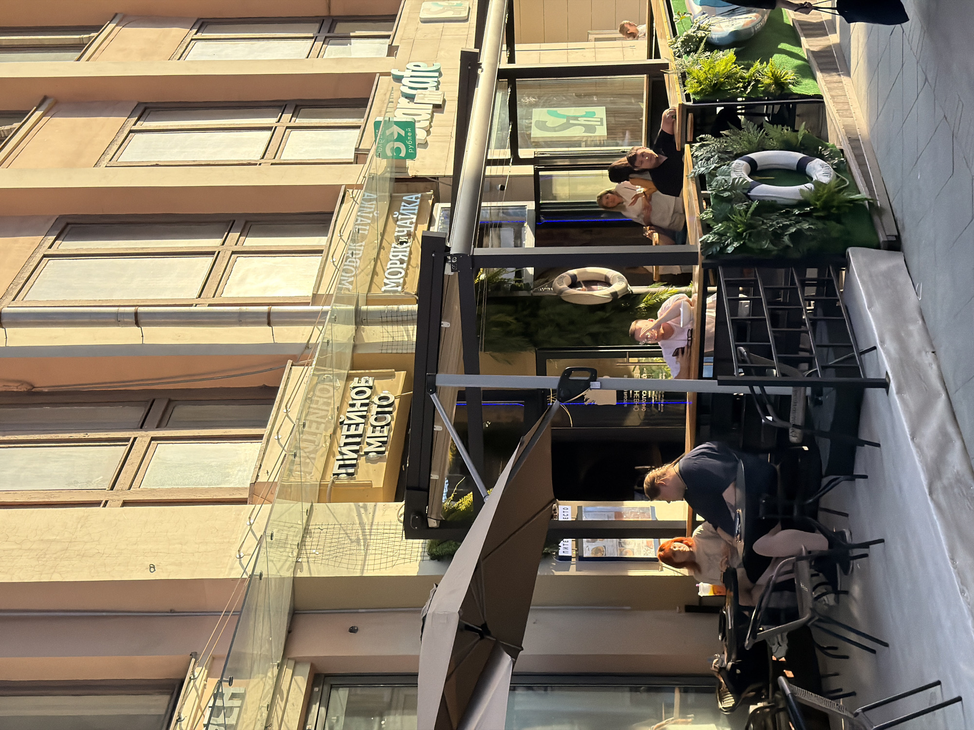 A daytime outdoor shot of the Питейное Место restaurant and бар 37 Cafe in Moscow, Russia. People are seated at tables. There is a large glass-covered area and several signs in Russian. A life preserver and plants decorate the outdoor space.