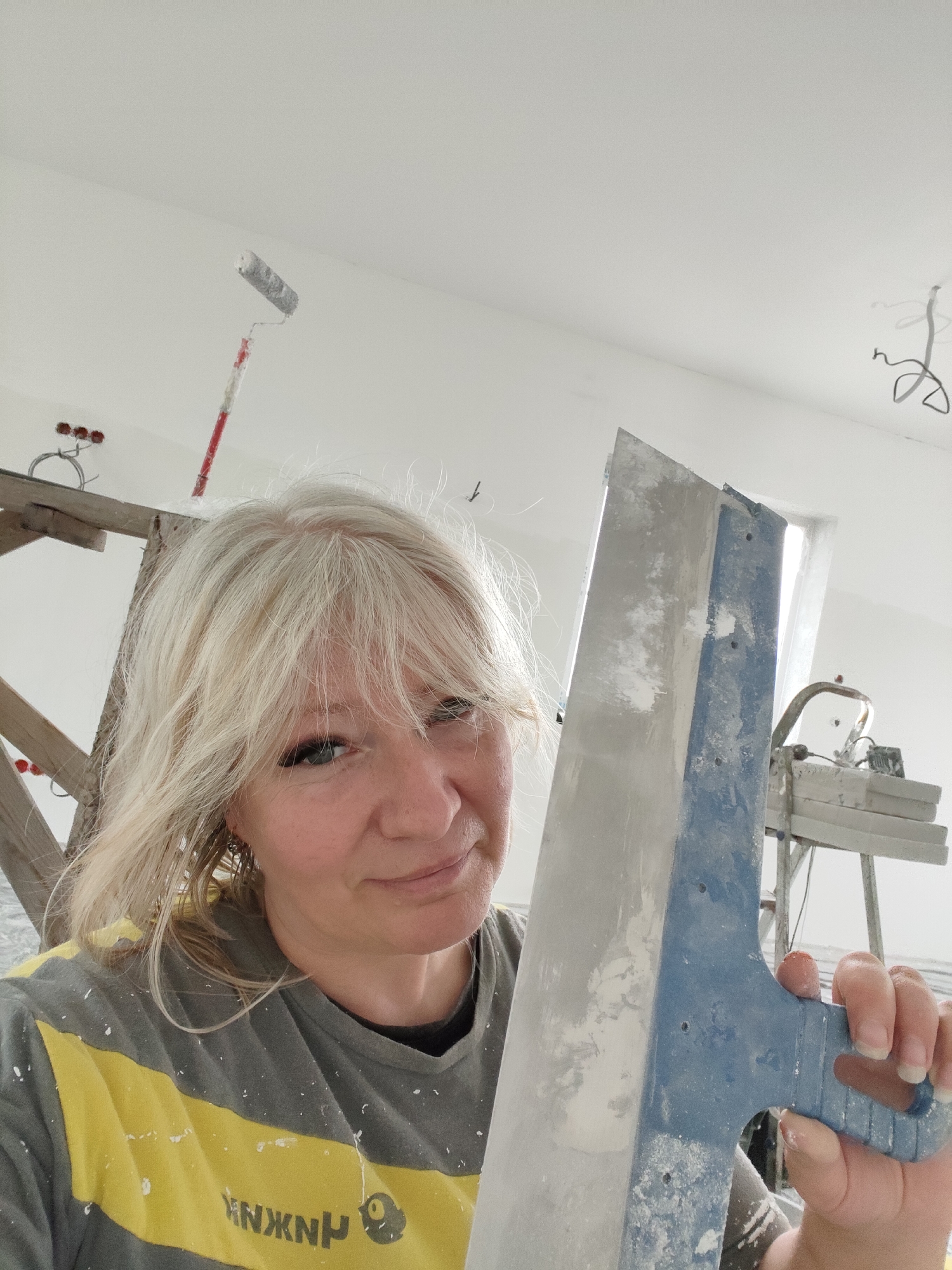 A woman with blonde hair is holding a large putty knife in what appears to be an unfinished room. The room has white walls and ceiling, and there are construction materials visible in the background, including a scaffold, a paint roller, and some electrical wiring. The woman is wearing a grey and yellow t-shirt, which is splattered with white paint or plaster. She is looking at the camera and smiling. The image appears to be a selfie, taken by the woman herself. The location is identified as Skurygino, Russia.