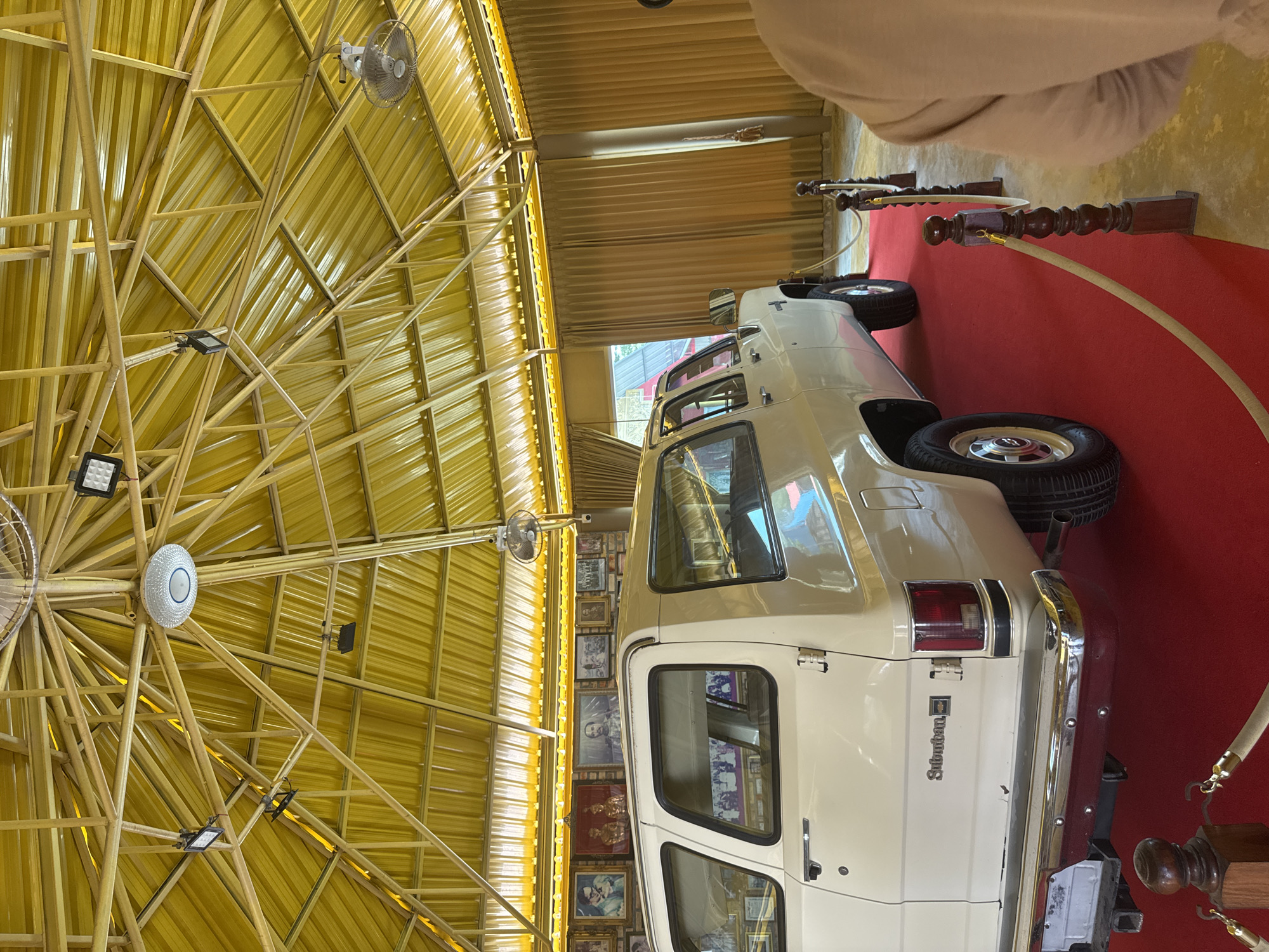 The image shows the interior of a building in Ban Khlong Khian, Thailand, featuring a yellow, radial-patterned metal roof structure with lights and fans. Below the roof, a cream-colored Chevrolet Suburban is on display, positioned on a red carpet. The Suburban's rear is prominently visible, with the "Suburban" nameplate and Chevrolet logo. The background wall is covered with framed photographs. A person is visible out of focus in the upper right corner of the image. Ornate wooden stanchions with ropes flank the red carpet.