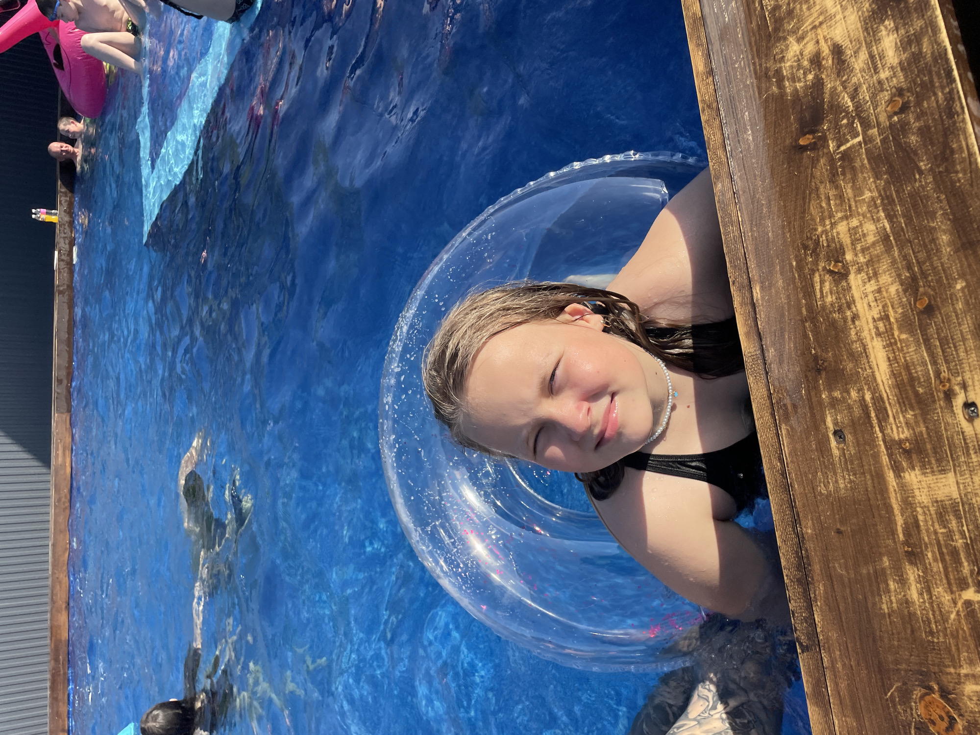 A girl is in a swimming pool using a clear inflatable tube. The pool is lined with wooden planks on one side and metal siding on another. There are other people swimming in the pool. The location is not discernible as Люкшудья, Russia without additional information.