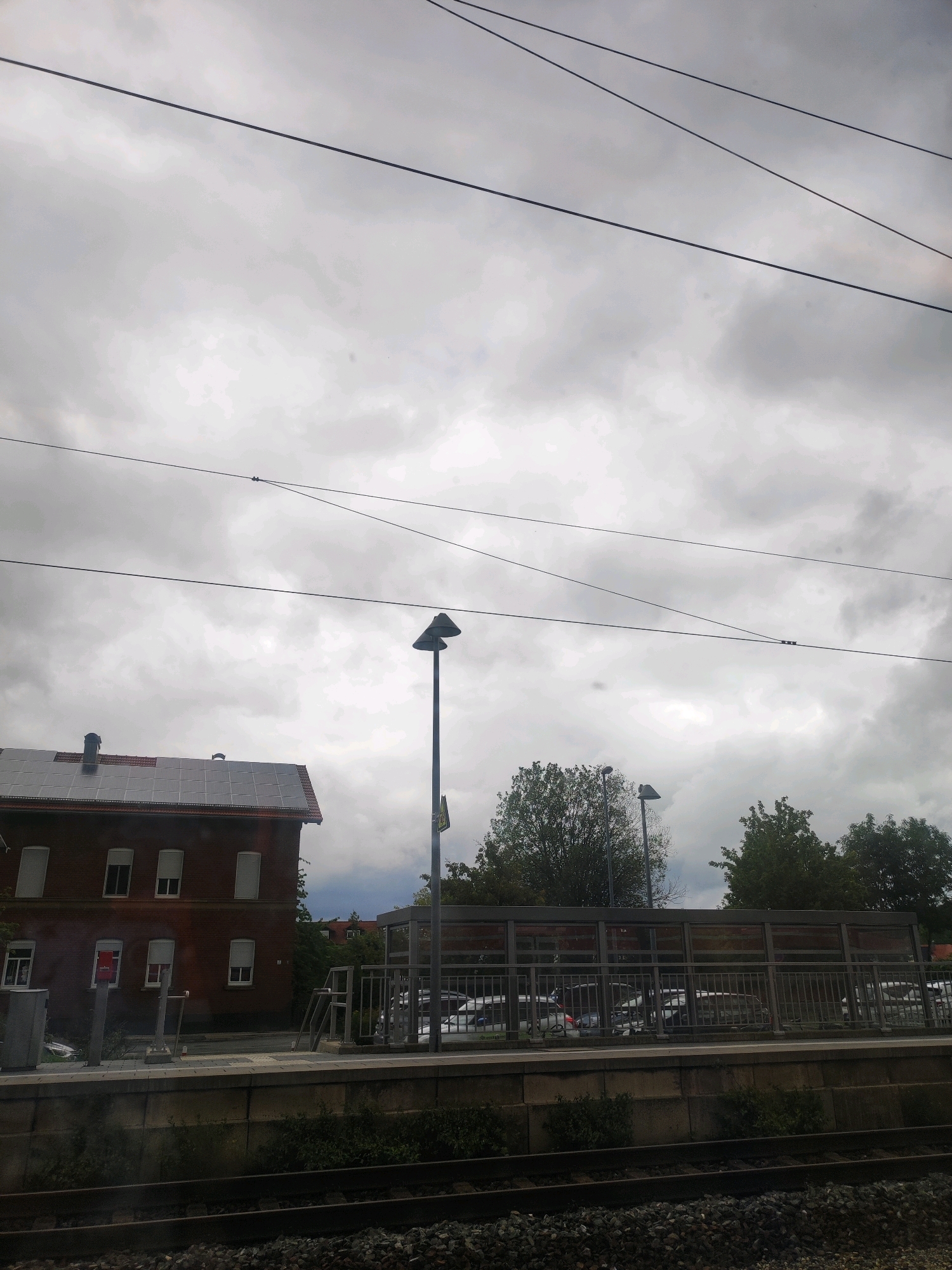 The image depicts a view from a train window, looking out at a cloudy sky and a scene in Petersaurach, Germany. In the foreground, train tracks are visible. Beyond the tracks is a raised platform, bordered by a concrete wall and greenery.
On the platform, a red brick building with solar panels on its roof is situated on the left. Several windows with white shutters are visible on the building's facade. To the right of the building, a lamppost with two lights stands tall. Further to the right, a covered waiting area with a glass facade and metal railings is visible, with several parked cars behind it. Trees are scattered throughout the scene, with some taller trees forming a backdrop. Overhead, multiple wires stretch across the frame, characteristic of a railway environment. No people are visible in the image.
