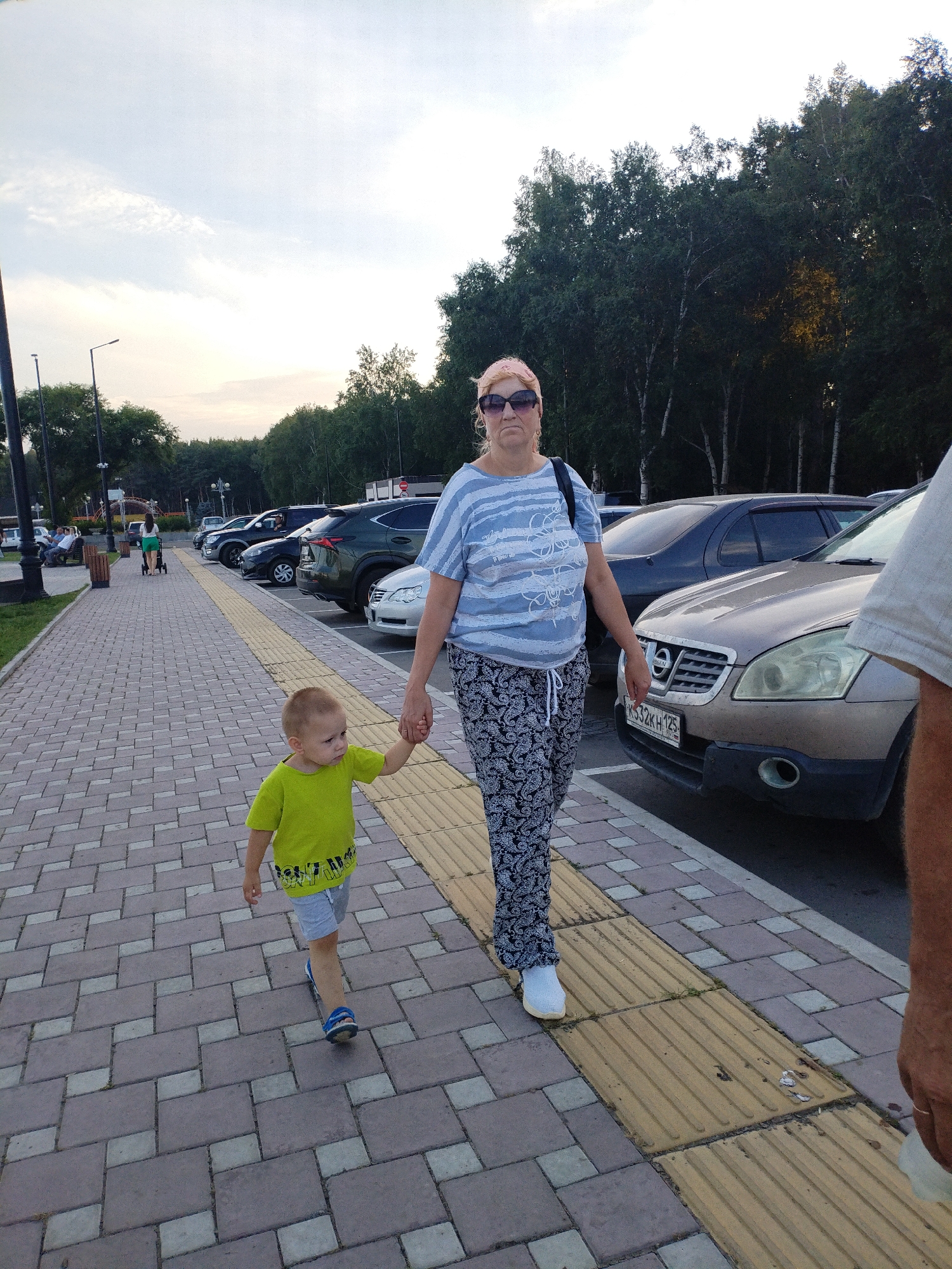 A woman and a young child are walking on a paved walkway in a city park. The woman, wearing a blue and white striped shirt and patterned pants, is holding the child's hand. The child is wearing a bright green shirt and gray shorts. In the background, trees and parked cars are visible. The scene appears to be a public area with a sidewalk, streetlights, and benches. This scene was captured on September 10, 2023, at 17:25:18 local time in Komsomolsk-on-Amur, Russia.