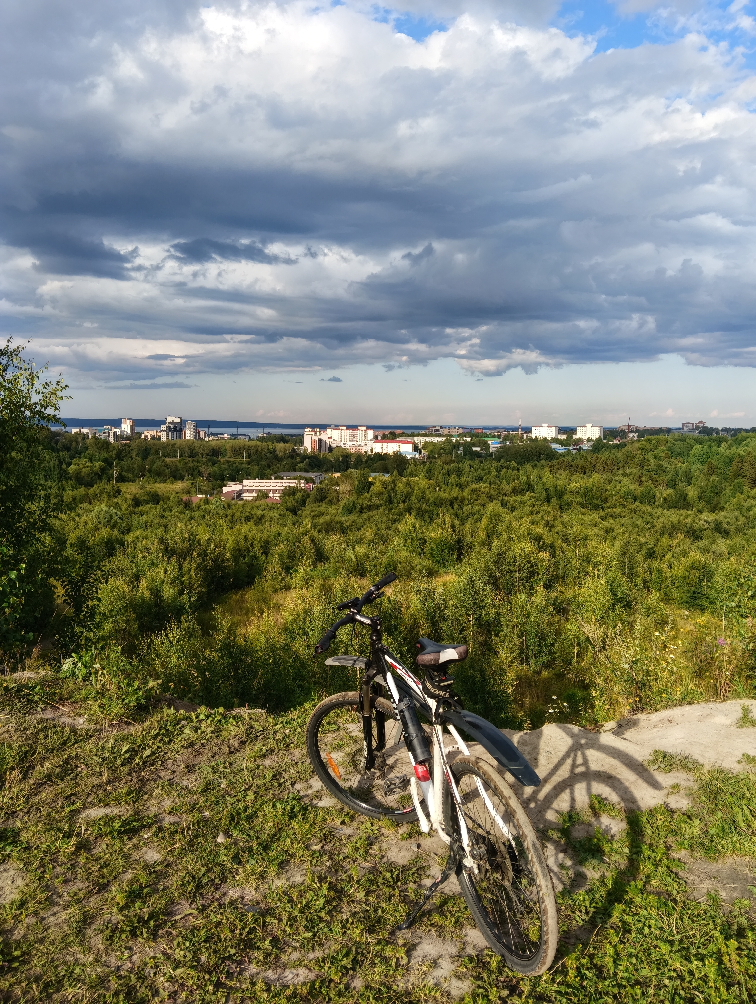 The image captures a scenic view from a high vantage point overlooking the city of Petrozavodsk, Russia. In the foreground, a white and black bicycle is parked on a grassy embankment. The bicycle has a fender on the rear wheel and a kickstand supporting it. Beyond the embankment, a vast expanse of dense green forest stretches towards the horizon. In the distance, a cityscape with various buildings, including multi-story residential structures and industrial elements, is visible. The skyline is bordered by a body of water, likely Lake Onega, and further beyond are more buildings and industrial structures. The sky is filled with dramatic, dark clouds interspersed with patches of bright blue sky, suggesting a dynamic weather system. The lighting indicates it is daytime, possibly late afternoon, given the shadows cast by the bicycle and trees. The overall impression is one of a natural landscape meeting an urban environment under a striking sky.