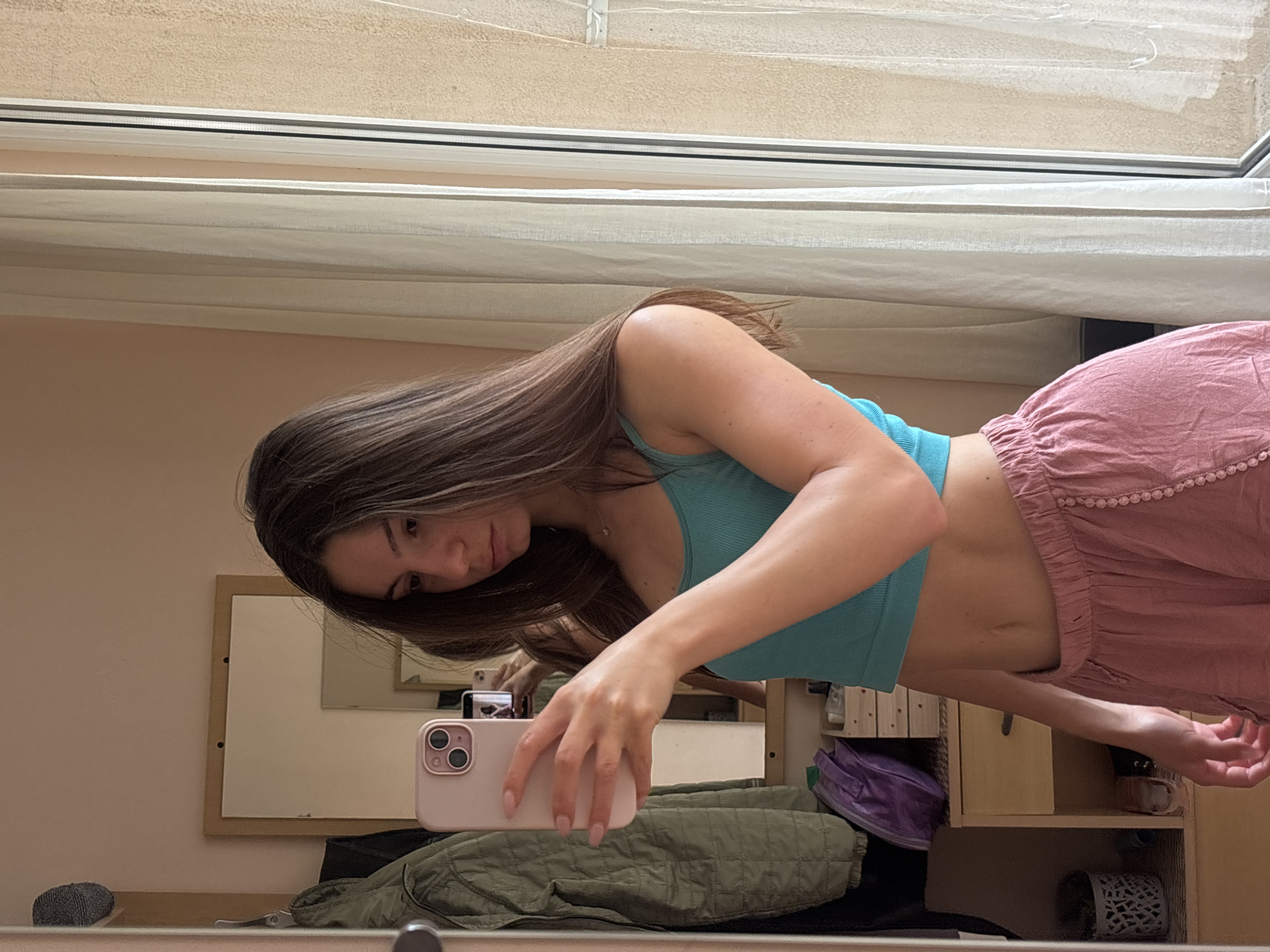 A young woman with long brown hair takes a mirror selfie in a room. She is wearing a turquoise tank top and pink shorts. Her left arm is raised, holding a pink smartphone up to take the photo. The background shows a mirror reflecting more of the room, including a quilted green jacket hanging on a hook, a small dresser with drawers, and a window with sheer curtains. A portion of what appears to be a building exterior is visible through the window. The scene suggests an indoor setting, likely a bedroom or dressing area. The location is not discernible from the image content.