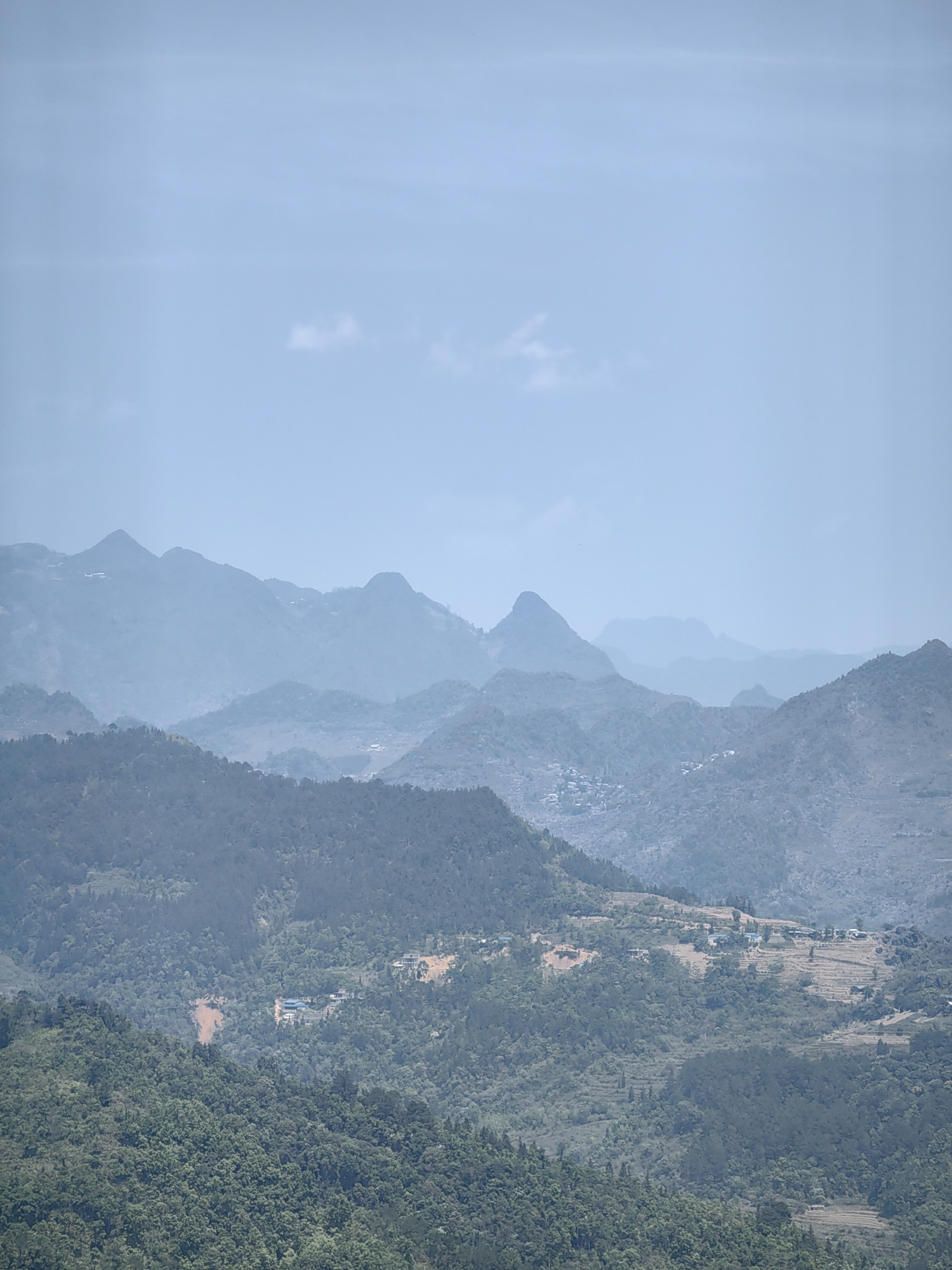 The image shows a hazy, distant view of mountainous terrain in Yên Minh Commune, Vietnam. Multiple layers of mountain ridges recede into the distance, becoming lighter in color due to atmospheric perspective. The foreground features lush, green forested hills with scattered settlements. The presence of clear-cut areas or terraced fields is visible on some of the hillsides. The sky is a pale blue with minimal cloud cover. The scene is rural with no apparent signs of urban development beyond the scattered settlements.