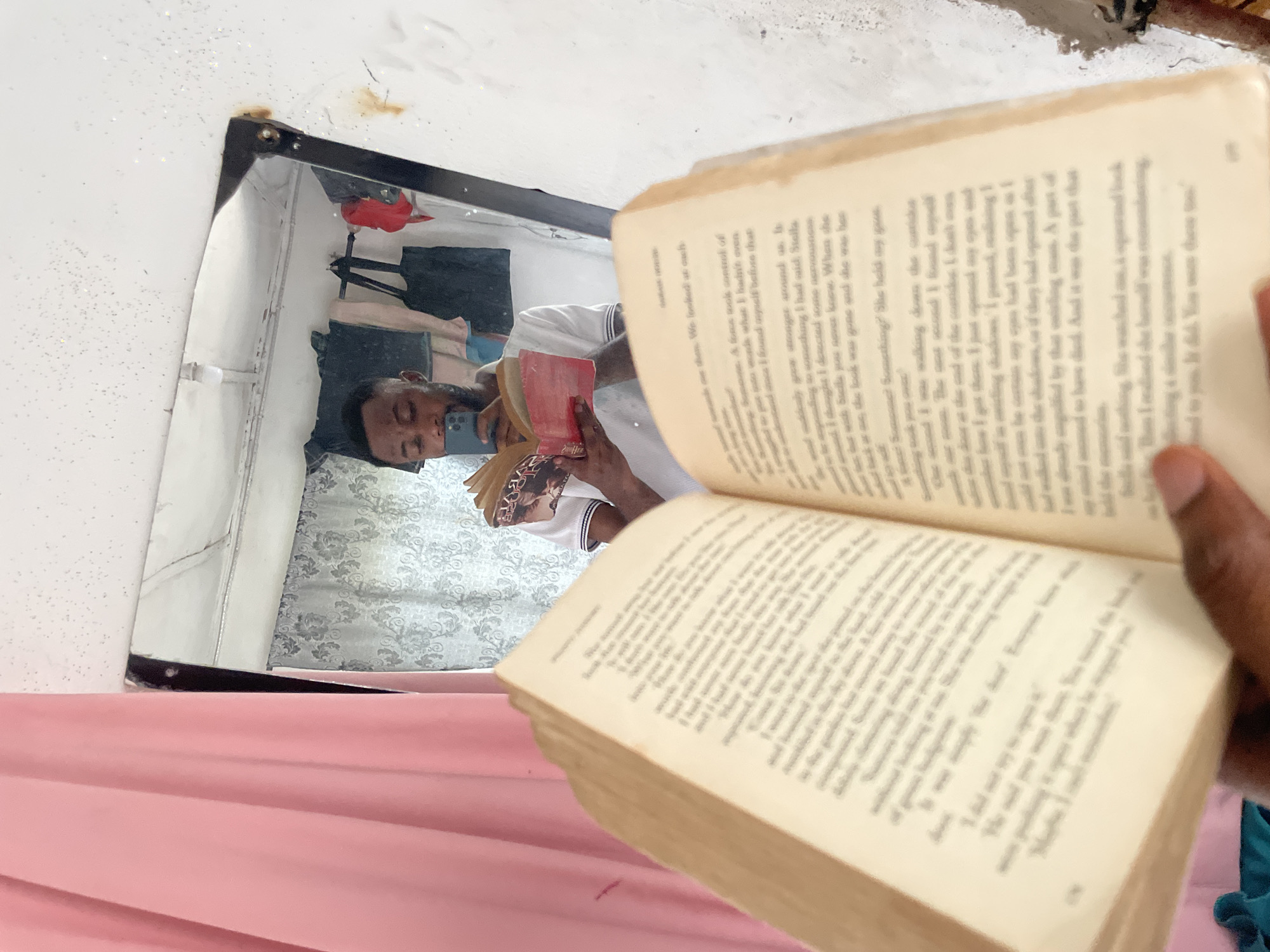 A man is reflected in a mirror while holding a book open in front of it. The man is wearing a white shirt with navy trim. He is holding a phone in his left hand and the book in his right. The mirror's reflection shows the man sitting on a bed with a gray and white patterned comforter. A pink fabric is draped in the foreground of the image. The open book is in focus, obscuring much of the reflection. The text on the pages is visible but unreadable. The mirror is framed in black and is mounted on a white wall. The top right corner of the mirror is slightly damaged. The location is not possible to confirm from the image.