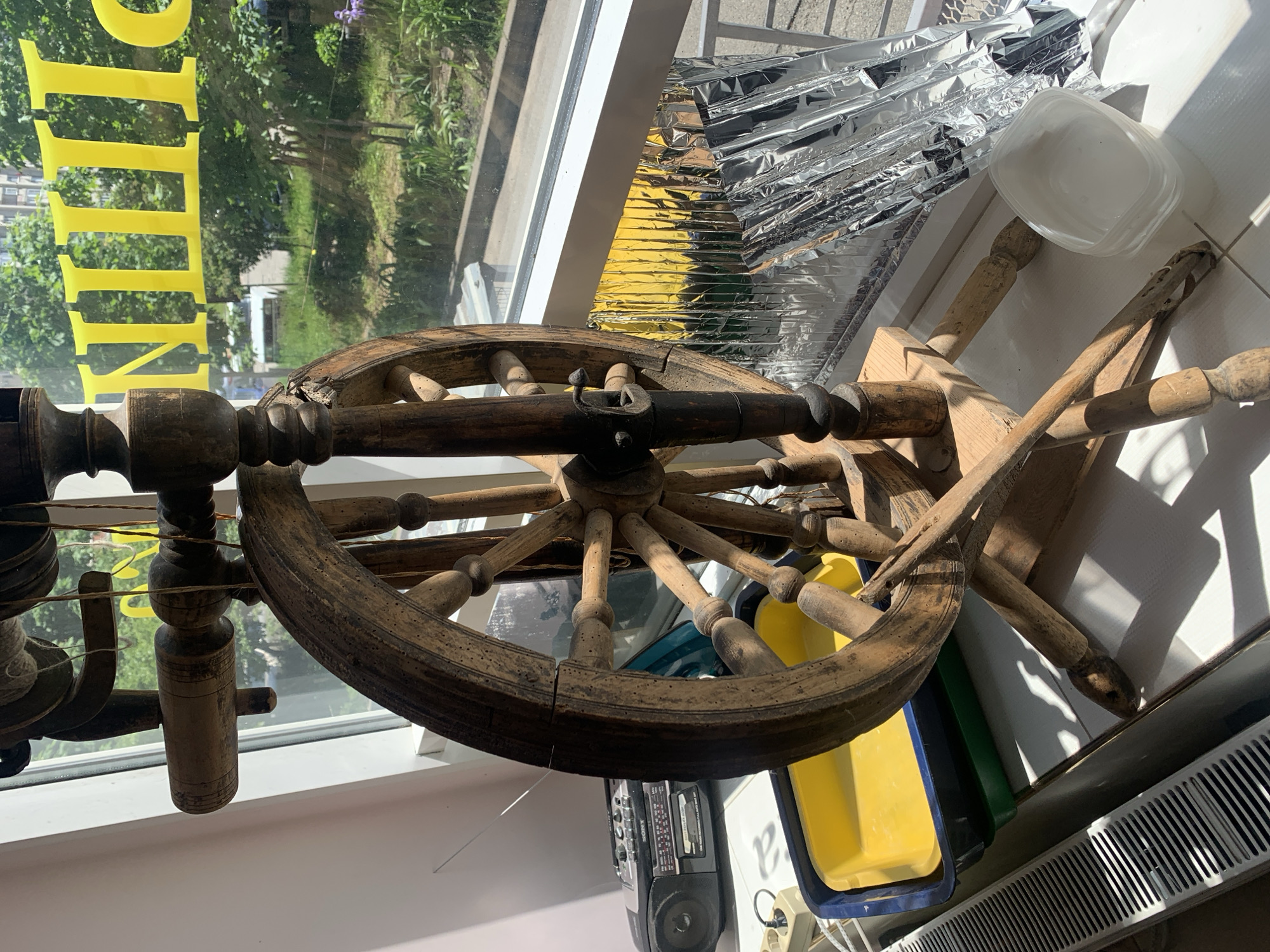 An antique-looking wooden spinning wheel is the central subject of this image, positioned indoors near a window. The spinning wheel, made of dark, worn wood, features a large spoked wheel, bobbins, and various carved components, with some string or yarn visible on one of the bobbins. The view through the window reveals green trees and foliage, suggesting an outdoor setting like a garden or park, possibly with distant buildings.
Bright sunlight streams through the window, indicating it is daytime and sunny. On the window, reflective yellow text is visible, partially spelling out what appears to be "MILLIN" or a similar word, possibly part of a business sign.
To the right and below the spinning wheel, several other objects are cluttered near the window. These include crumpled reflective foil material, possibly used for insulation or sun reflection, stacked clear plastic containers, and yellow and blue plastic bins. Further down, a black portable radio or boombox (with the "Sony" brand partially visible) sits on a surface. A white radiator is also visible along the bottom edge of the frame. The overall scene depicts a somewhat cluttered indoor space with a mix of functional and decorative items, illuminated by natural light. The location is Stavropol, Russia.