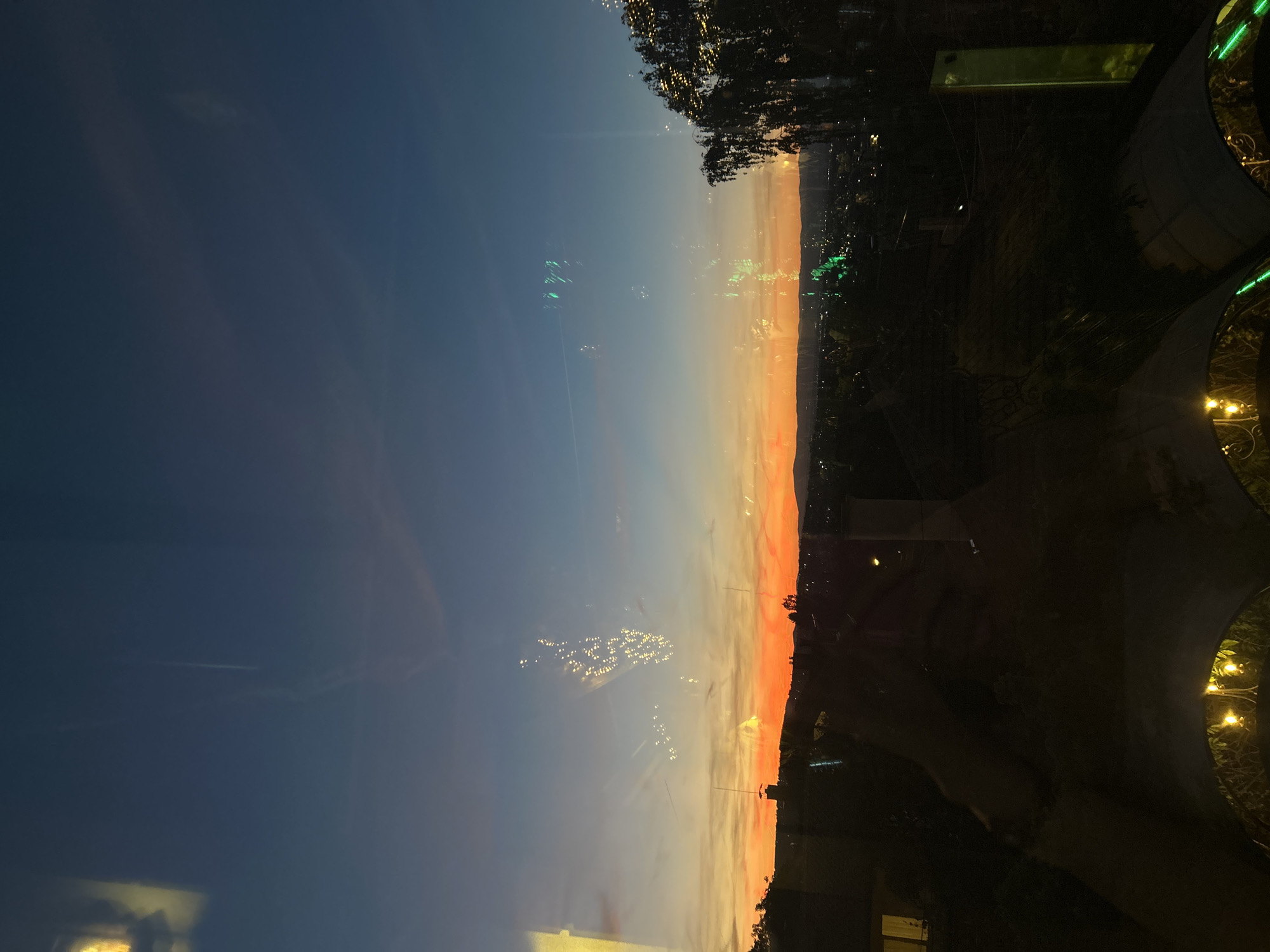The image depicts a sunset sky transitioning from blue to orange. The lower portion shows a dark silhouette of land or buildings, with sparse lighting indicating an urban environment. The reflection of lights suggests observation from inside a building, possibly in the city of Sofia, Bulgaria. Specific identifiable landmarks or people are not visible.