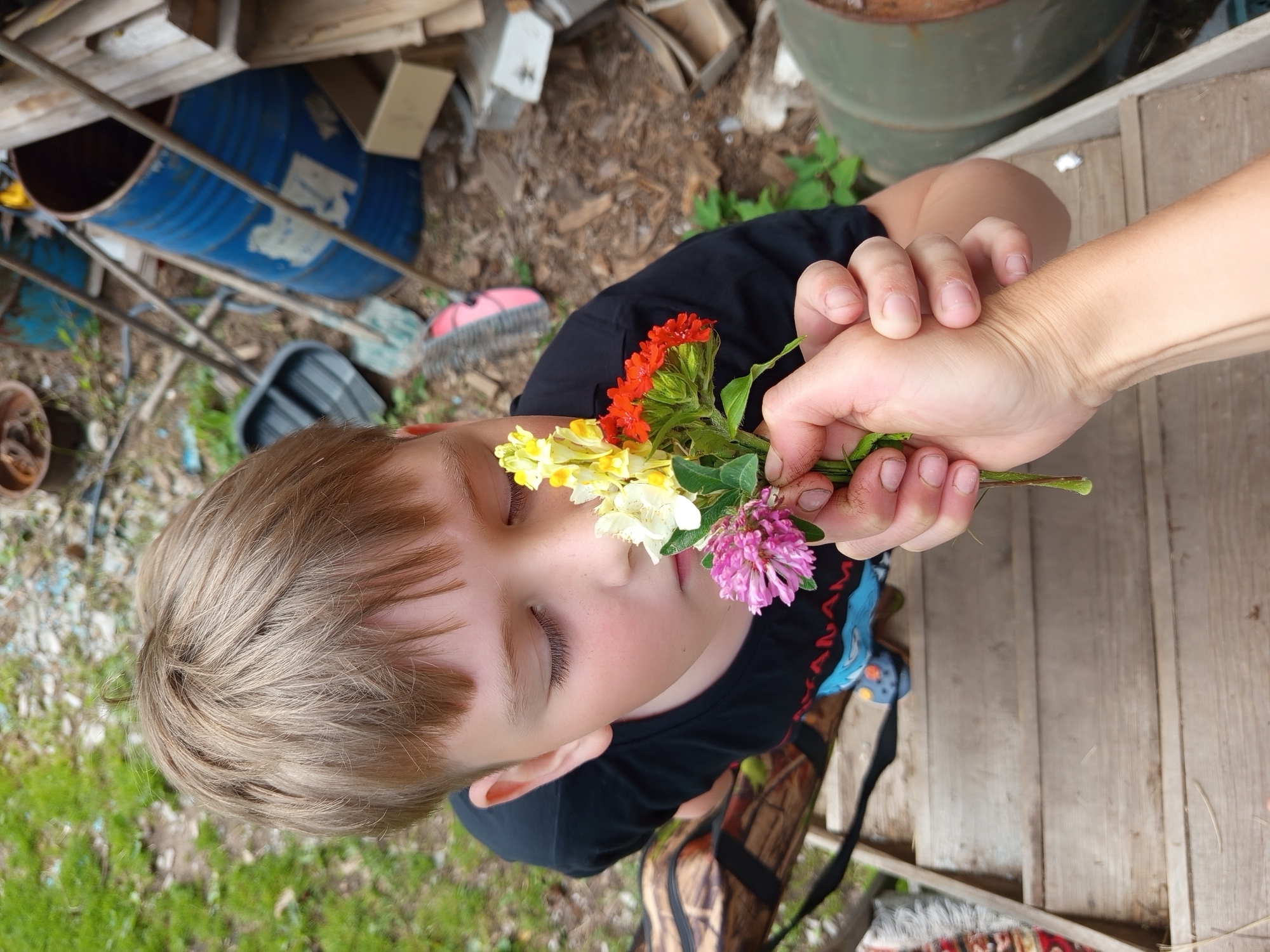A young boy with blond hair and closed eyes is presented with a small bouquet of wildflowers, including red, yellow, and pink blossoms. The boy wears a black t-shirt. A second person's hand is holding the bouquet to the boy's face. The scene appears to be outdoors, with a background of stacked wood, blue barrels, and dry ground mixed with green grass. Wooden planks are visible in the foreground. The image has a candid feel, suggesting a casual moment, possibly during daylight. There is no visible text. The overall impression is one of innocence and a simple connection with nature.