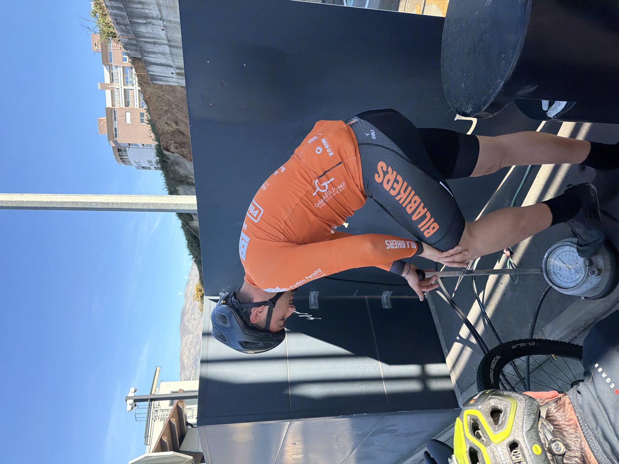 Here's a detailed description of the image:
**Subject and Actions:** The image shows a male cyclist dressed in an orange and black "BILA BIKERS" uniform, including a black cycling helmet. He is bending over, seemingly inflating the tire of a bicycle. He appears to be using a gauge to measure the tire pressure.
**Environment:** The photo appears to be taken outdoors, possibly at an event or race staging area. In the background, there's a building structure with windows, suggesting an urban setting. The presence of a clear blue sky indicates a sunny day.
**Objects:**
* Bicycle and parts (tire, wheel)
* Air pressure gauge
* Helmet (partially visible, different from the one worn by the cyclist)
**Location:** Based on the text on the cyclist's uniform "Porfirio Pereira RICAS", "GAMAFRIGMF", and "BILA BIKERS", it is likely that this is a cyclist competing in a race located in Vila Real, Portugal.