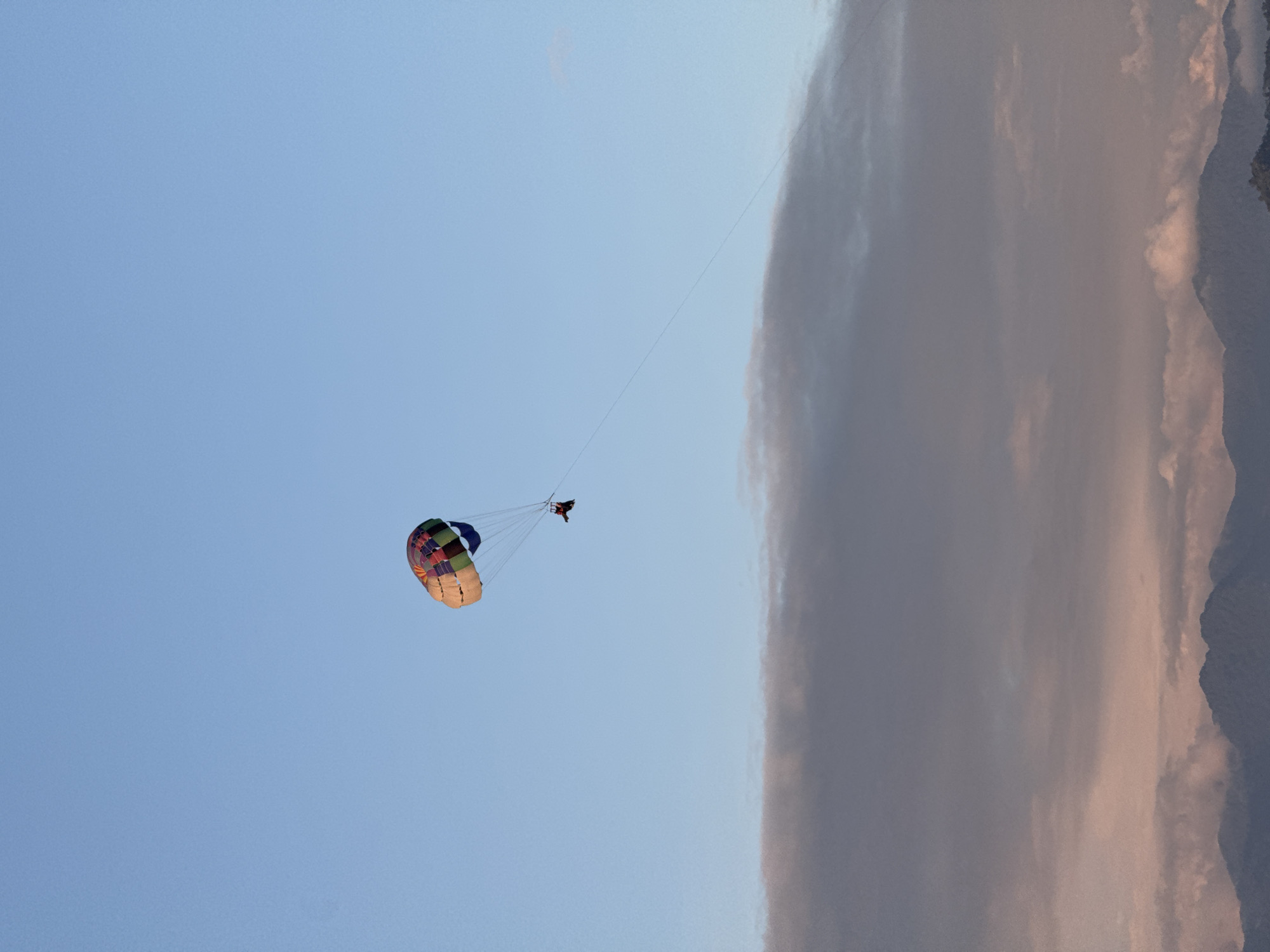The scene depicts a parasailer suspended in the air against a backdrop of a clear, light blue sky on the left and a large, vertical bank of gray clouds and mountains on the right. The parasail is multicolored with sections of blue, green, red, and yellow and has a canopy shape. A single person is hanging below the parasail. The parasail and the person are connected by multiple lines. A thin rope extends from the top of the parasail to the right edge of the frame, suggesting it is connected to a boat out of view. The setting appears to be Batumi, Georgia, based on the combination of the coastal environment, mountains, and parasailing activity, a common tourist attraction in the city.
