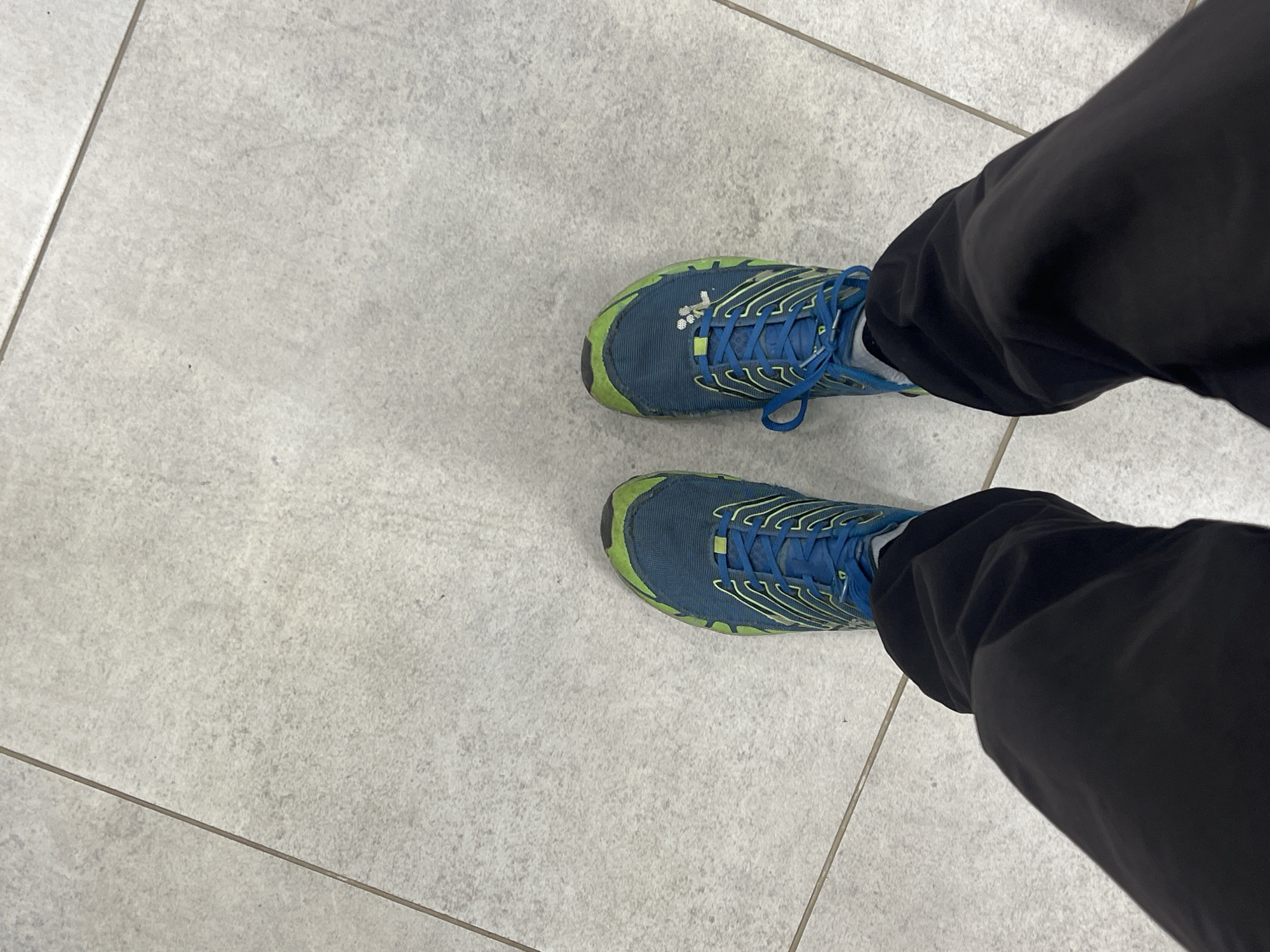 The image depicts a first-person view looking down at a pair of blue and green athletic shoes on a tiled floor. The lower portion of a person wearing black pants is visible on the right side of the frame. The background consists of light gray square floor tiles with dark grout lines. The exact location within the city of Perm, Russia cannot be determined from the image alone.