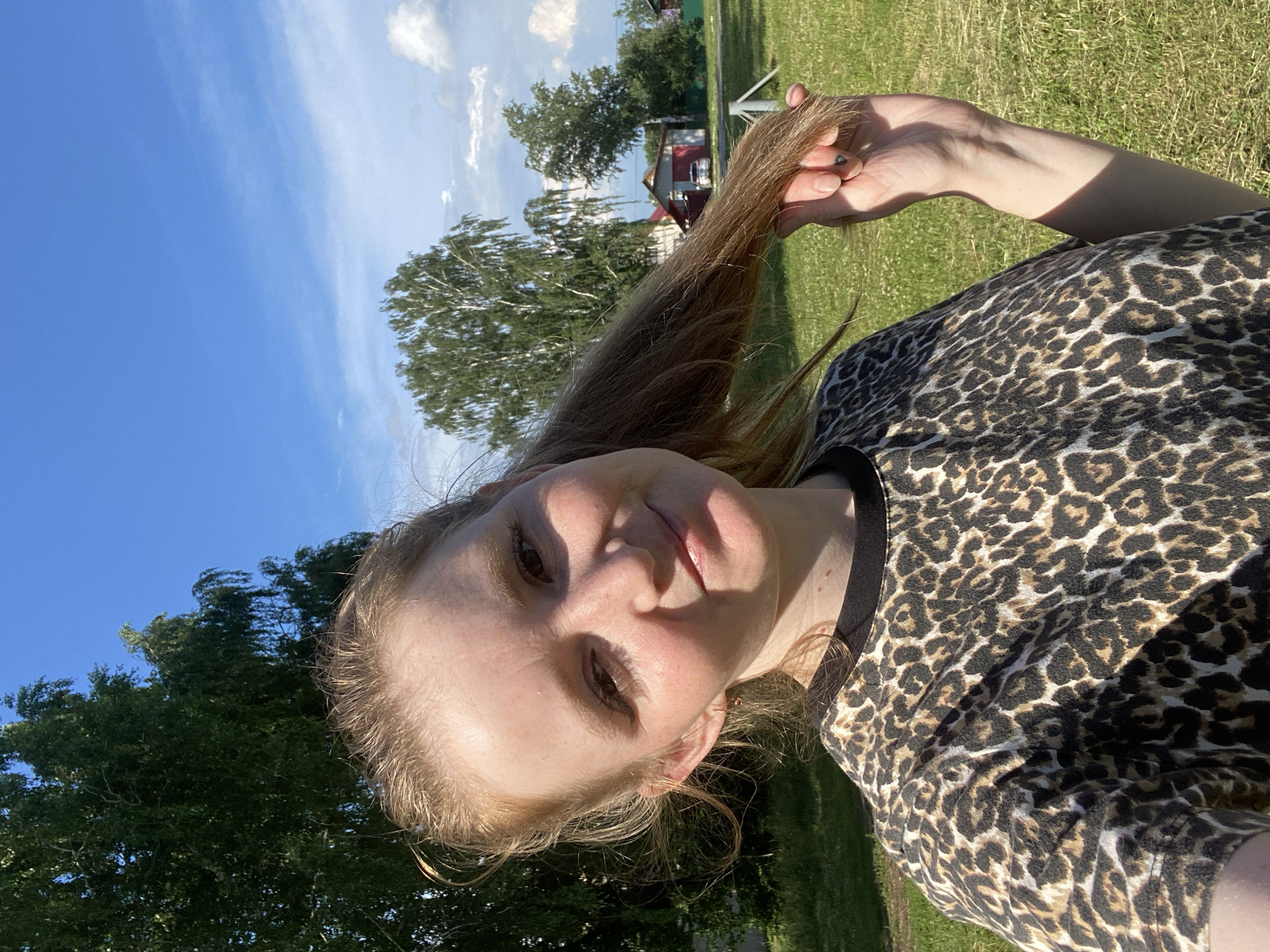 A young Caucasian woman with blonde hair tied in a ponytail is taking a selfie outdoors. She is wearing a black and brown leopard-print t-shirt and has a slight smile on her face. Her left hand is raised, holding a portion of her hair. The background shows a bright blue sky with scattered clouds, green trees, and a few buildings, suggesting a rural or suburban setting. The woman's facial features are soft, with freckles visible on her cheeks and nose. The lighting indicates it is daytime, likely sunny. The environment appears to be a grassy field. The buildings in the background seem to be houses with red roofs.