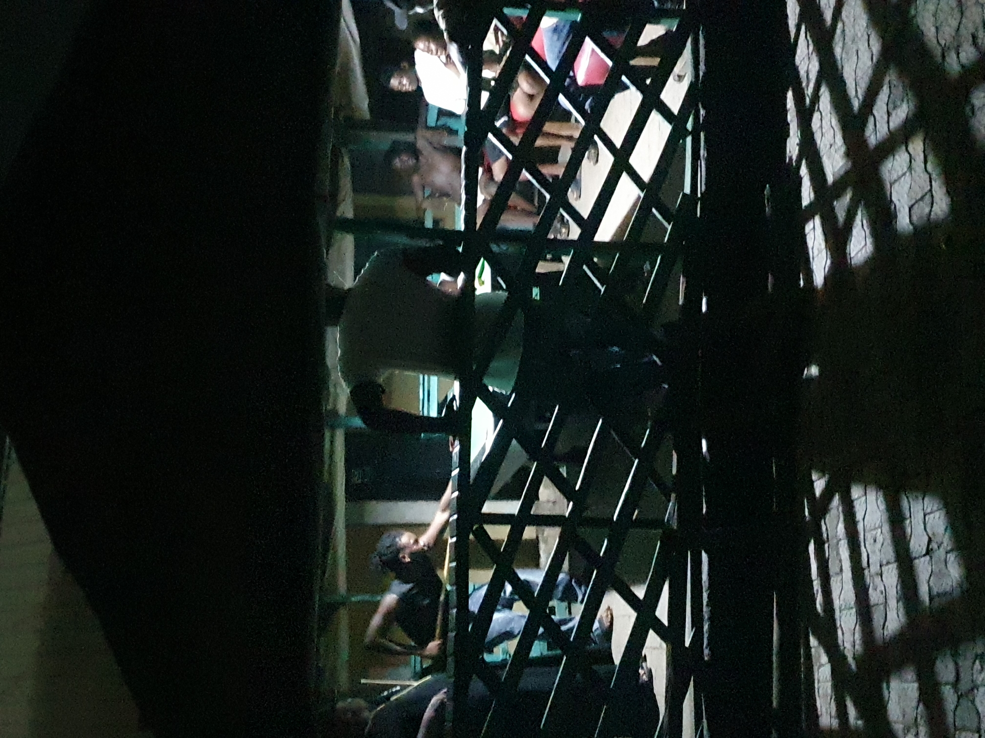 In Lokoja, Nigeria, a lattice gate partially obscures a group of people in a poorly lit interior. A man in a white shirt and dark pants is partially visible through the gate, as is another man wearing a dark shirt who appears to be climbing the structure behind it. Other figures are visible behind the gate, some in lighter clothing. The surrounding area is predominantly dark, with shadows cast by the gate onto what appears to be a wall.