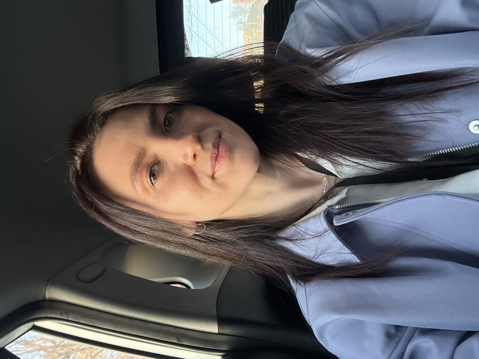 A self-portrait captures a woman seated inside a car, likely in Kazan, Russia. The image is oriented with a 90-degree counter-clockwise rotation. The woman, with long, dark brown hair styled with a side part, has brown eyes and a neutral to slightly soft expression. She wears a light periwinkle or light blue zippered jacket, featuring a snap button, over a white inner layer. A delicate silver chain necklace and a small silver hoop earring on her left ear are visible.
She is positioned in what appears to be the passenger seat of the vehicle, which has dark interior elements including the seat and the ceiling headliner. Through the car window, bare tree branches and a bright sky are discernible, indicating daylight hours and potentially a colder season. Bright sunlight illuminates her face and hair. Visible car interior details include the dark grey headliner and a window frame. There is no visible text in the image.