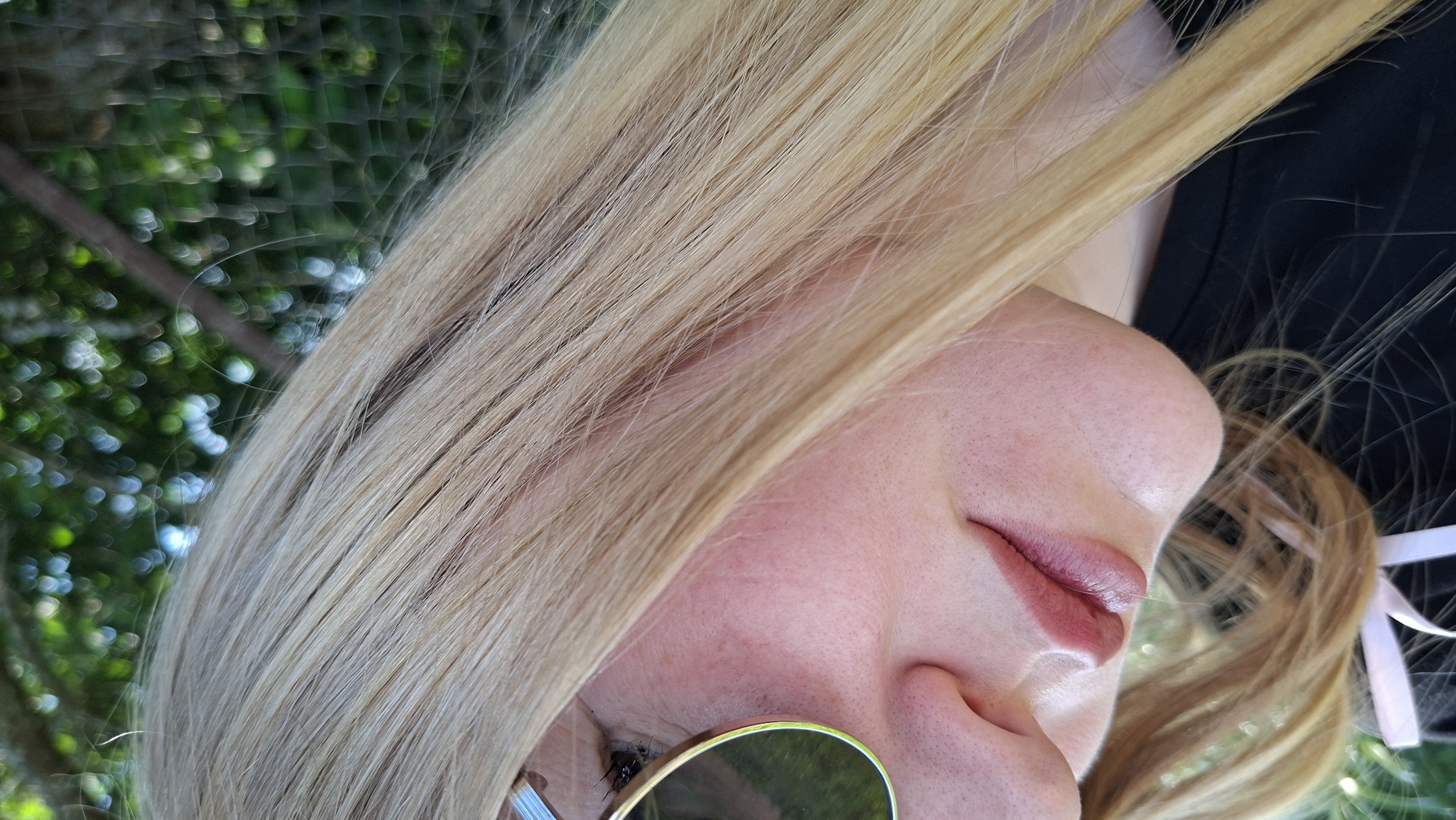 The image is a close-up, angled shot of a person's face and blonde hair, with a blurry green and dark background that appears to be foliage and a fence. The person is wearing sunglasses with round, reflective lenses and a dark top. The skin has a pinkish hue and visible pores. The lips are slightly parted and appear to have a subtle pink lipstick. A white ribbon is visible in the lower portion of the image. The specific location in the city of Большой Букор, Russia, cannot be determined from the visual information provided in the image.