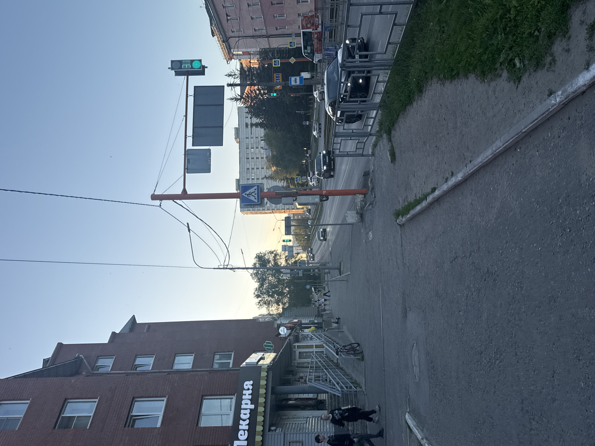 The image depicts a street scene in Barnaul, Russia, likely during late afternoon or early evening due to the lighting. A multi-story brick building, identified by the signage "Пекарня" (Bakery), is visible on the left. The building has multiple windows and an entrance with a ramp and stairs. Sidewalks are paved, with a portion showing exposed gravel. A metal fence separates the sidewalk from the road. The road is a multi-lane street with vehicles in motion. A traffic light with a green signal is visible, as are traffic signs. Several pedestrians are walking on the sidewalk. Trees are present along the roadside, and a tall apartment building can be seen in the background. The sky is clear and blue.