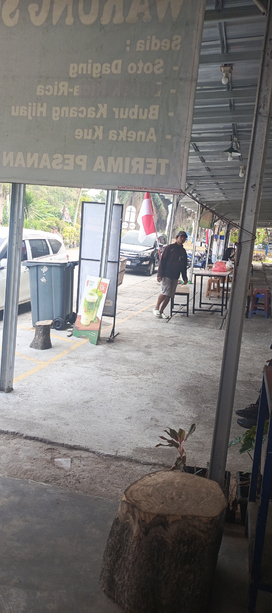 The image shows an outdoor seating area with a concrete floor and a covered roof. In the foreground, there is a large tree stump serving as a seat. To the left, a gray garbage bin with wheels is visible. A sign displaying a drink with "10K" price is propped up on an easel. A man wearing a brown jacket and shorts stands near a table with chairs. In the background, cars are parked, and a red and white Indonesian flag is displayed. The overall scene suggests a casual eatery or roadside stall. The sign in the background appears to be a menu with items like "Soto Daging" and "Aneka Kue". The sign also contains the text "TERIMA PESANAN" which translates to "Orders Accepted" in Indonesian. The location is identified as Sukaresmi, Indonesia.