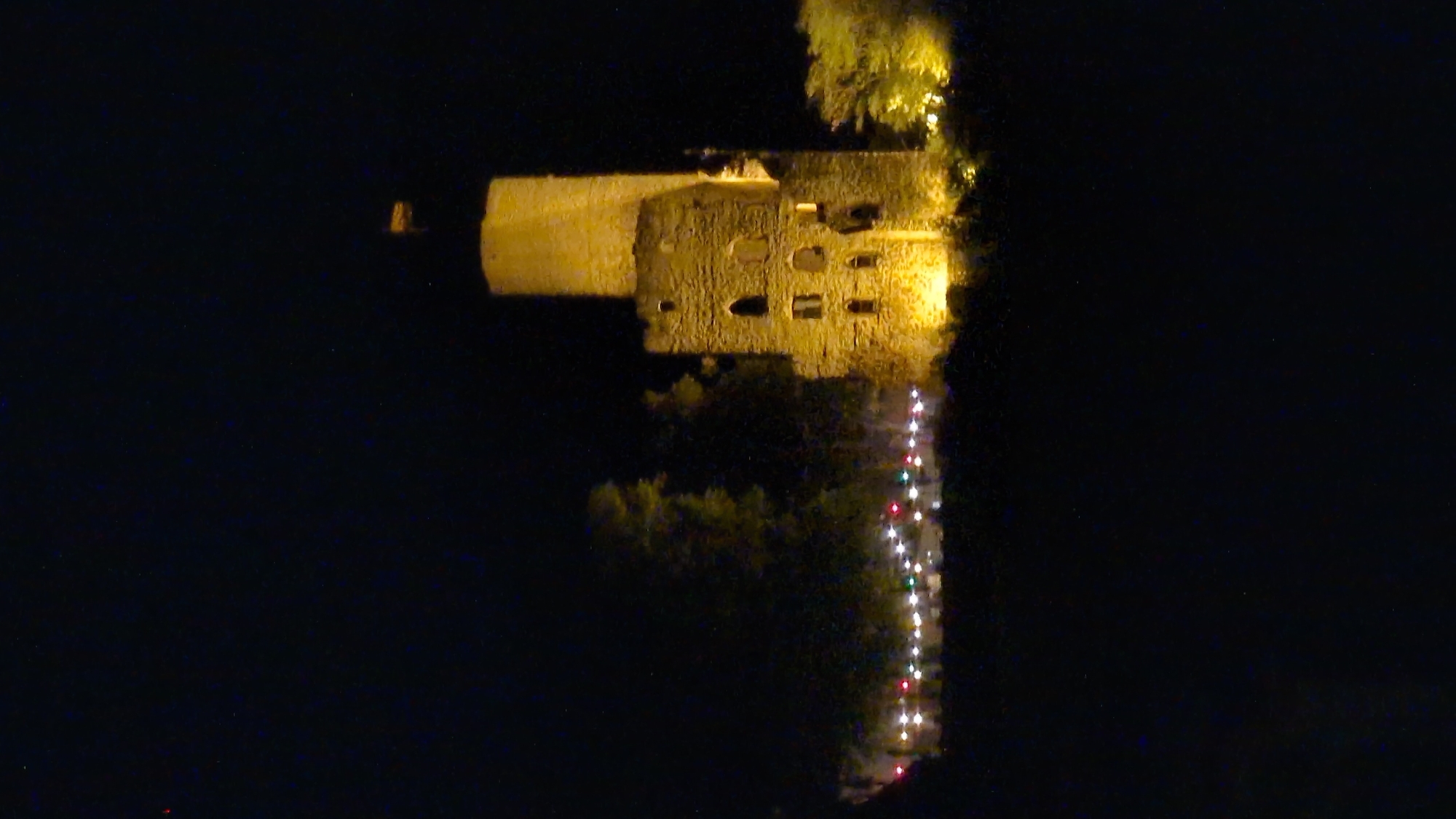 A medieval stone tower ruin, illuminated by artificial lights, stands tall against a black night sky. The tower has a cylindrical upper section and a more fragmented lower section with visible windows. In front of the tower, a line of colorful string lights is visible, suggesting an event or decoration. Trees and foliage surround the base of the tower, also partially lit. The scene is set at the Strahlenburg castle in Schriesheim, Germany, which is a landmark and ruin overlooking the town. There are no people or discernible activity depicted in the image.