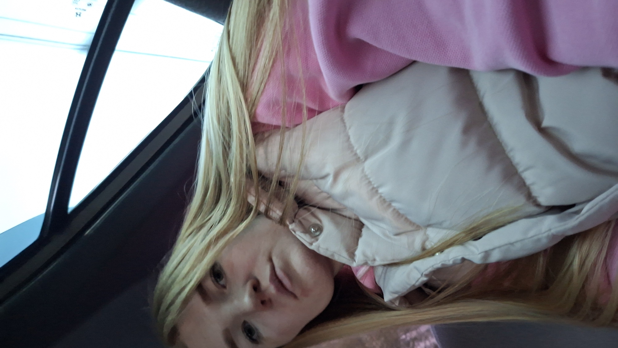 A blonde woman with light skin is wearing a pink sweater and a beige puffer vest inside a vehicle. She is positioned near the window, which is partially open. The location is not identifiable as Chaikovsky, Russia, based on visual elements alone.