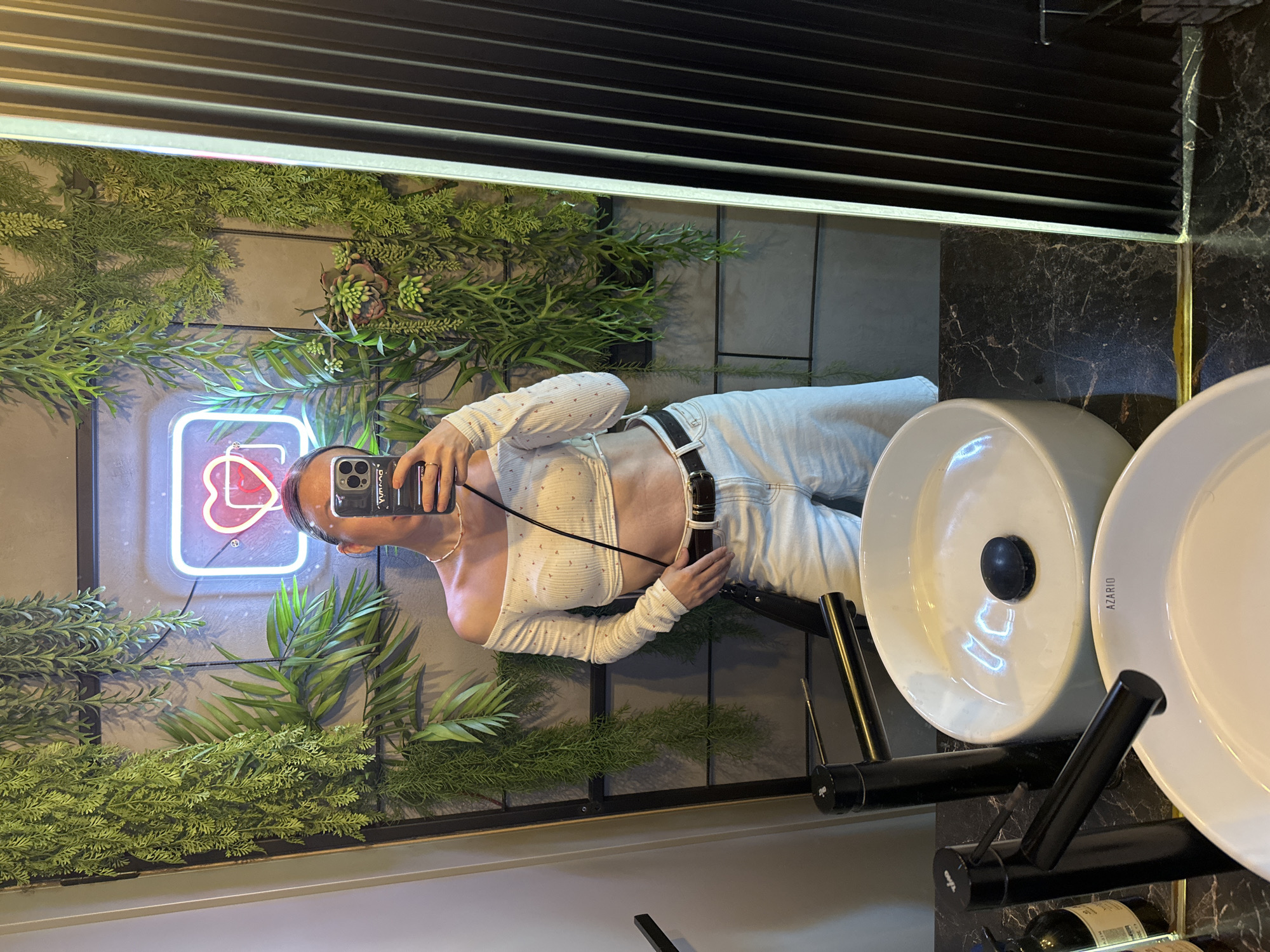 The image is a mirror selfie taken in a bathroom. The reflection shows a person holding a smartphone, dressed in a white, long-sleeved crop top and light-colored jeans with a black belt. The bathroom decor includes lush green plants and a neon sign with a heart shape. Two white washbasins are visible, one with a black drain plug. The background shows tiled walls and a black, slatted structure above the mirror. The location in Votkinsk, Russia is not discernible from the image.