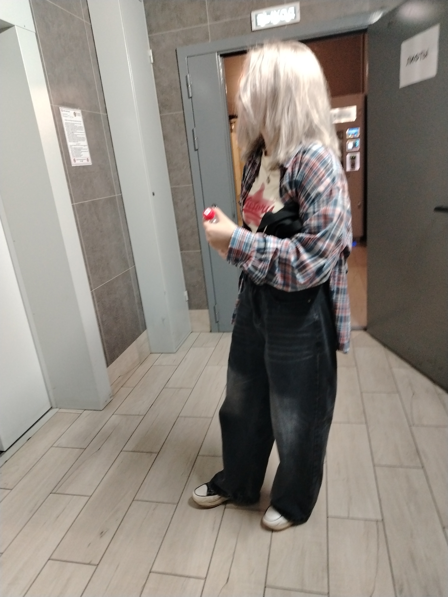 A person with shoulder-length blonde hair is standing in a hallway, facing away from the camera and towards an open elevator doorway in Izhevsk, Russia. They are wearing a black t-shirt, a red and blue plaid flannel shirt, and baggy dark-wash jeans with white sneakers. They are holding a small red object in their right hand. The hallway has light-colored tile flooring and gray tiled walls. There are two elevator banks, and signage is visible above one of the elevator doors, and on the wall to the right of the door in Russian.
